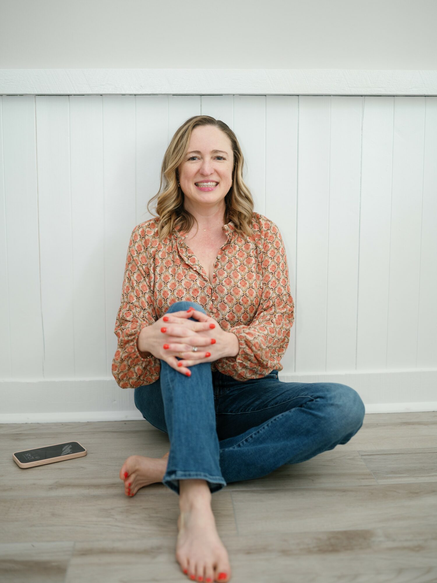 Woman sitting on floor with crossed legs, smiling, holding a phone, against white wall background.