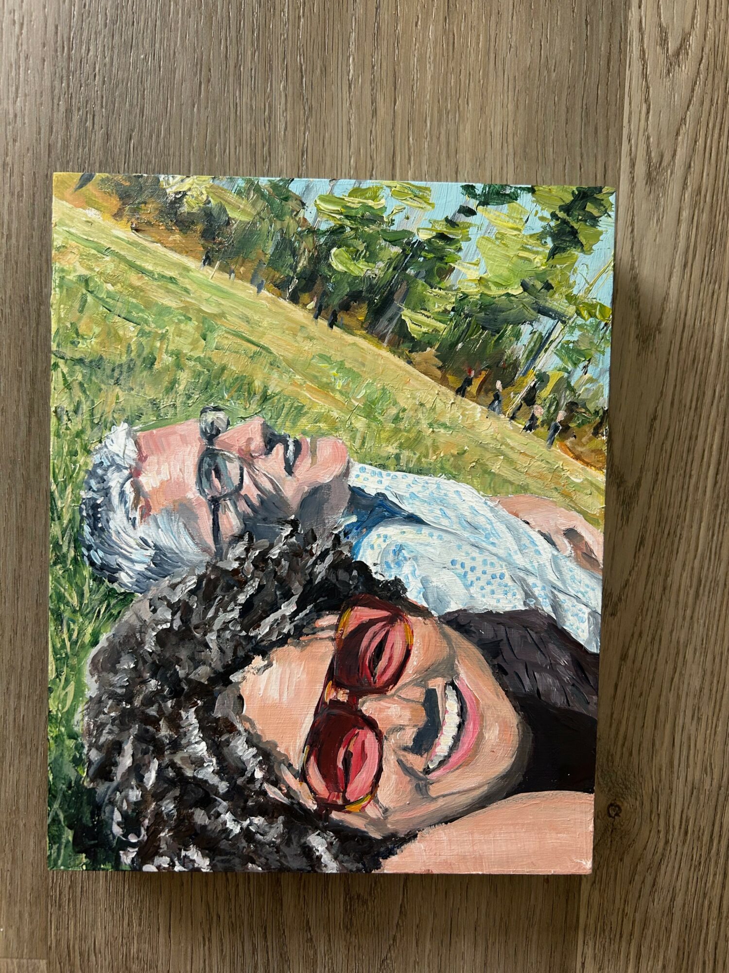 Person with curly hair and sunglasses smiling outdoors, lying on grass with trees and a blue sky in background.
