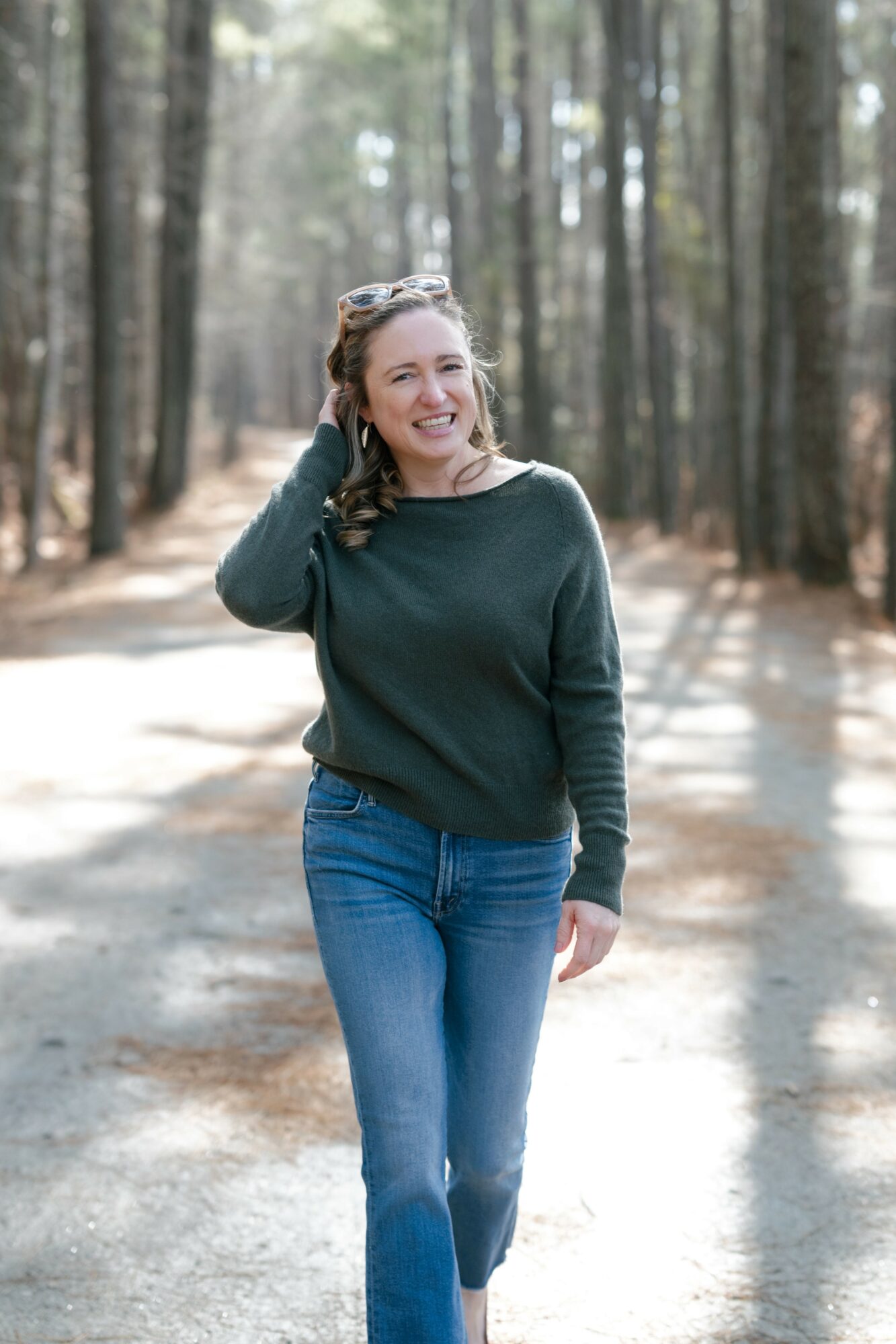 Woman walking in a forest, smiling, wearing a dark sweater and jeans, sunlight filtering through trees.