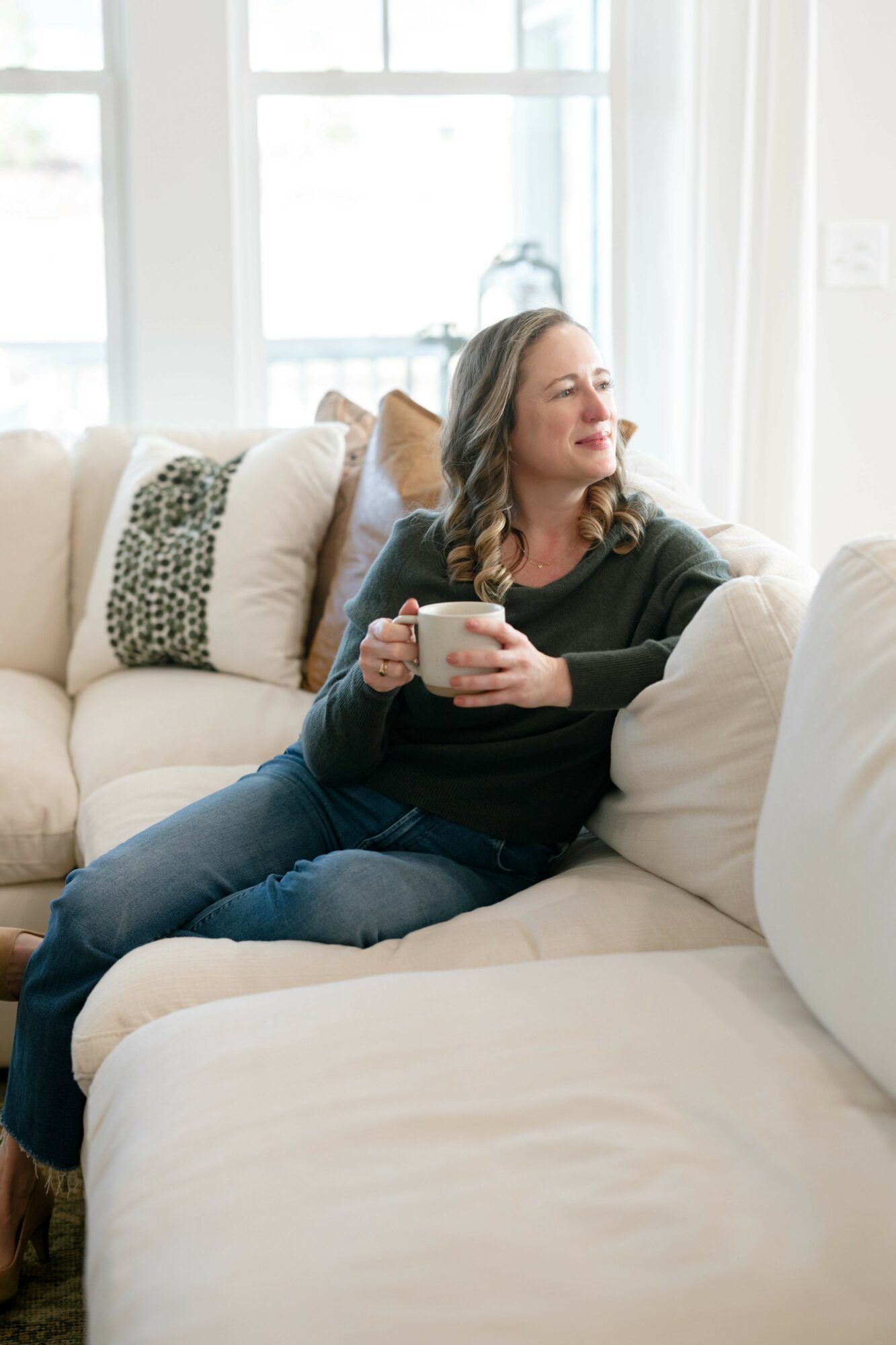 Woman sitting on a sofa holding a mug, smiling with eyes closed, in a bright living room.