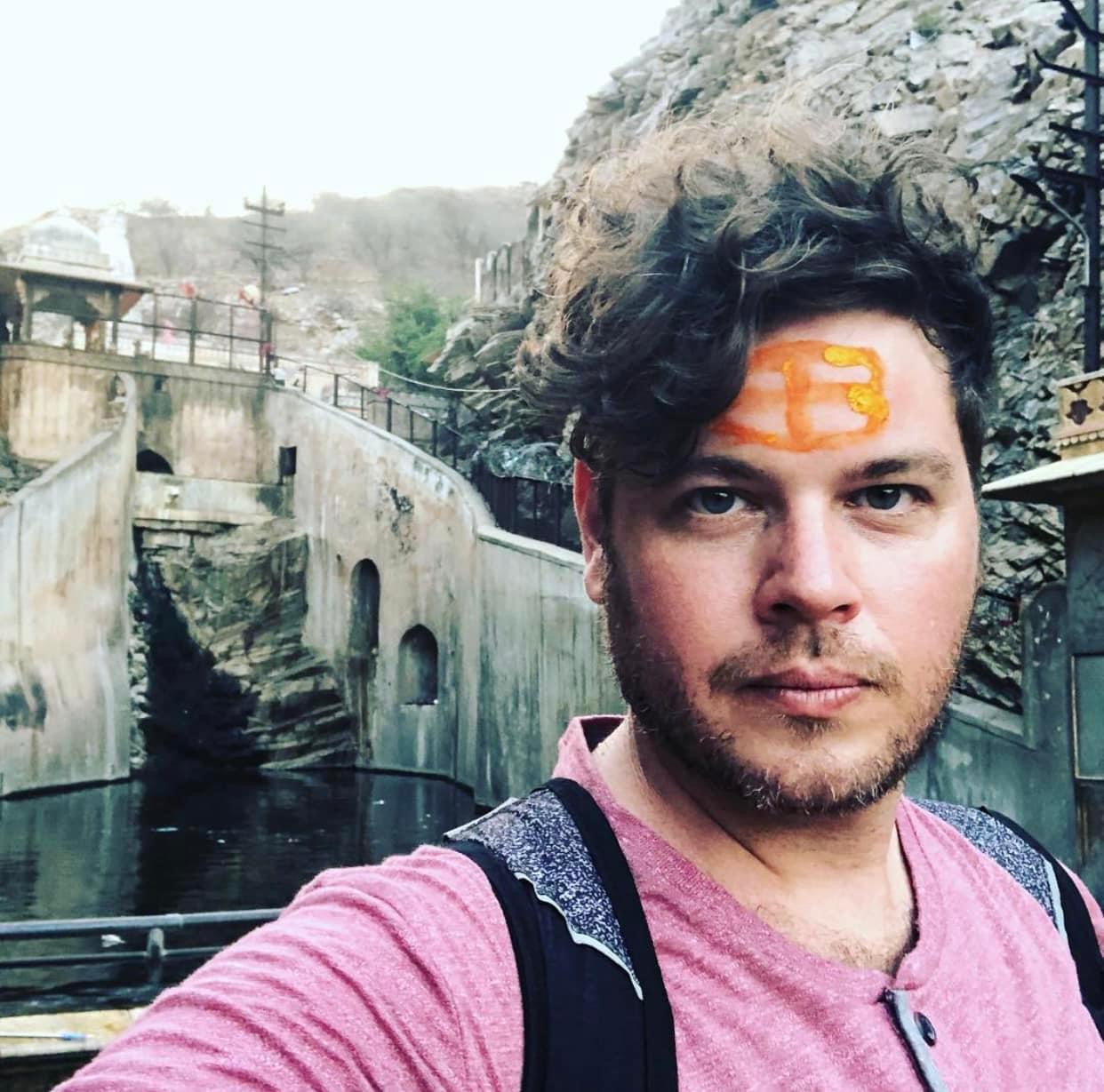 Young man with curly hair and face paint, wearing a pink shirt and backpack, outdoors near rocky terrain.