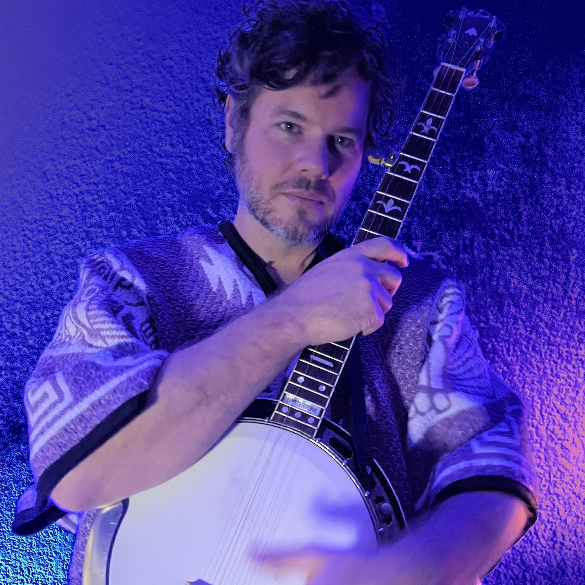 Man holding a banjo, looking at the camera, against a textured wall, illuminated with purple and blue lighting.