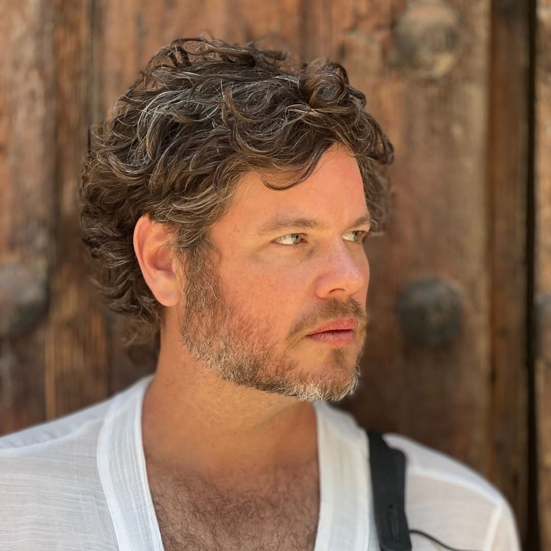 Man with curly hair and beard looking to the right, standing in front of a wooden background.