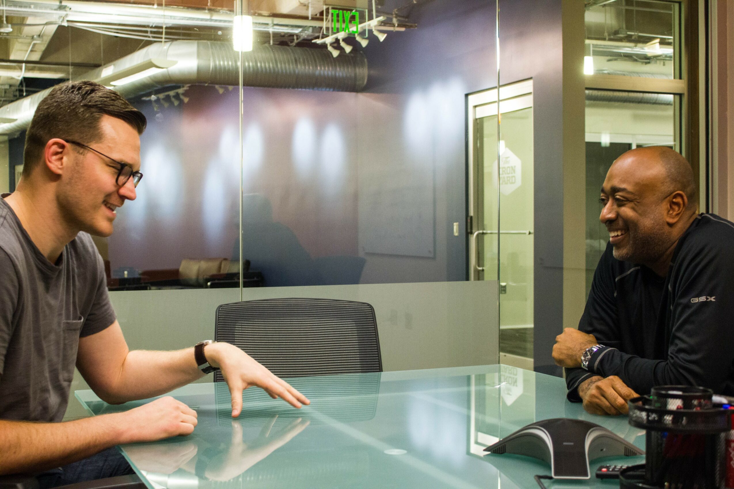 Two men sitting at a glass table, smiling and talking in a modern office setting.