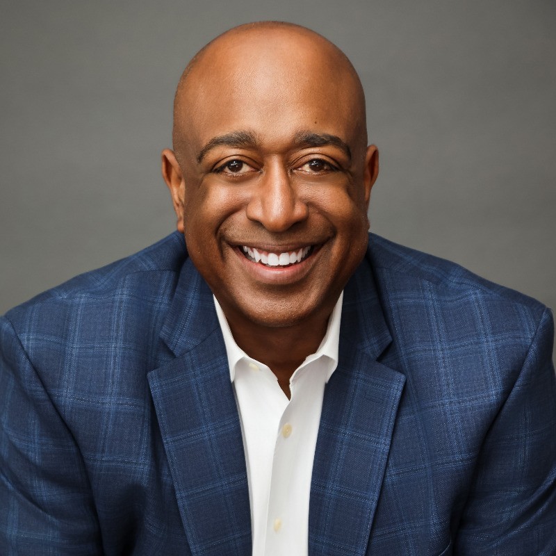 Smiling man with a bald head wearing a blue checkered suit and white shirt against a gray background.