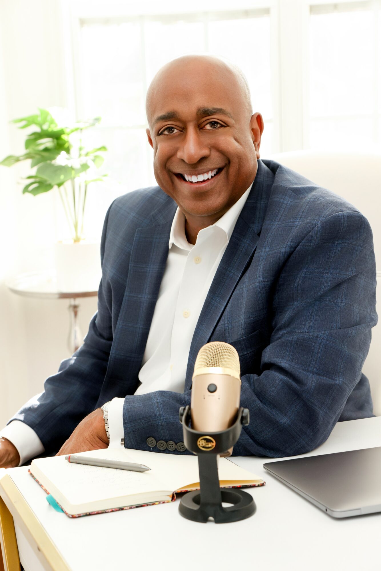 Man in suit smiling at desk with microphone, notebook, and laptop, in bright room with window and plant.