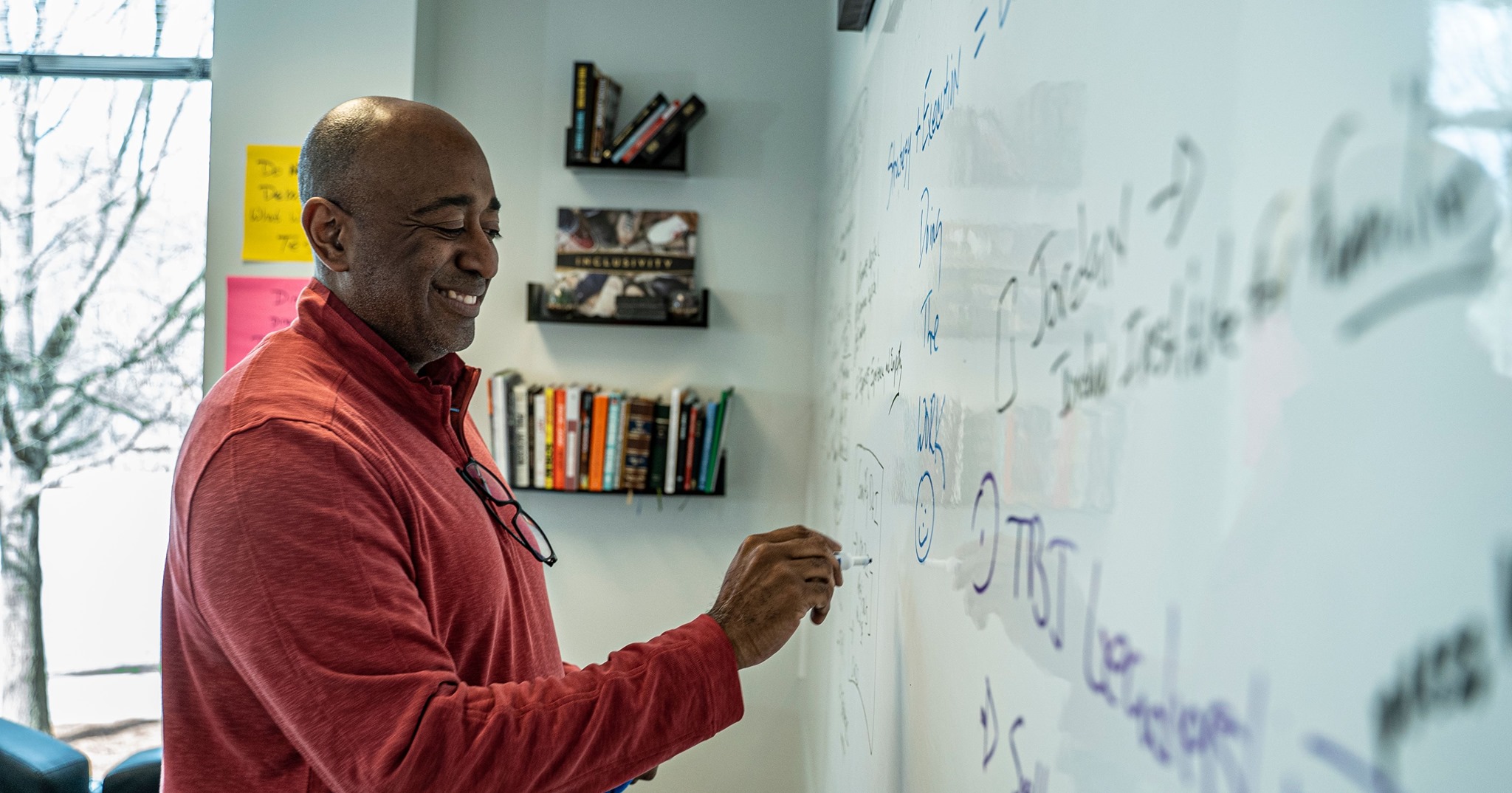 Man writing on a whiteboard in a room with bookshelves, smiling, with a window in the background.