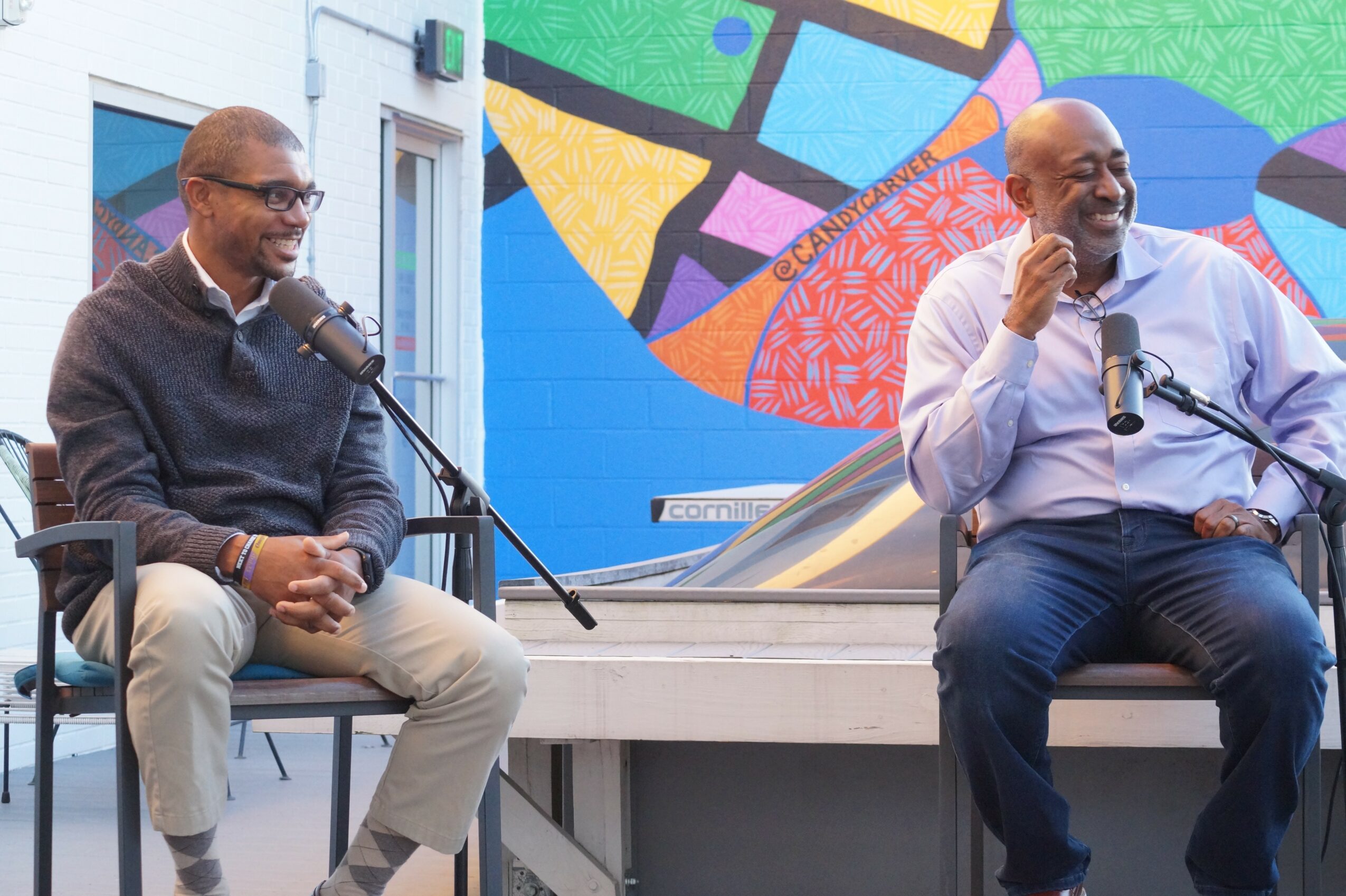 Two men sit on a stage with microphones, engaging in conversation, with a colorful mural in the background.