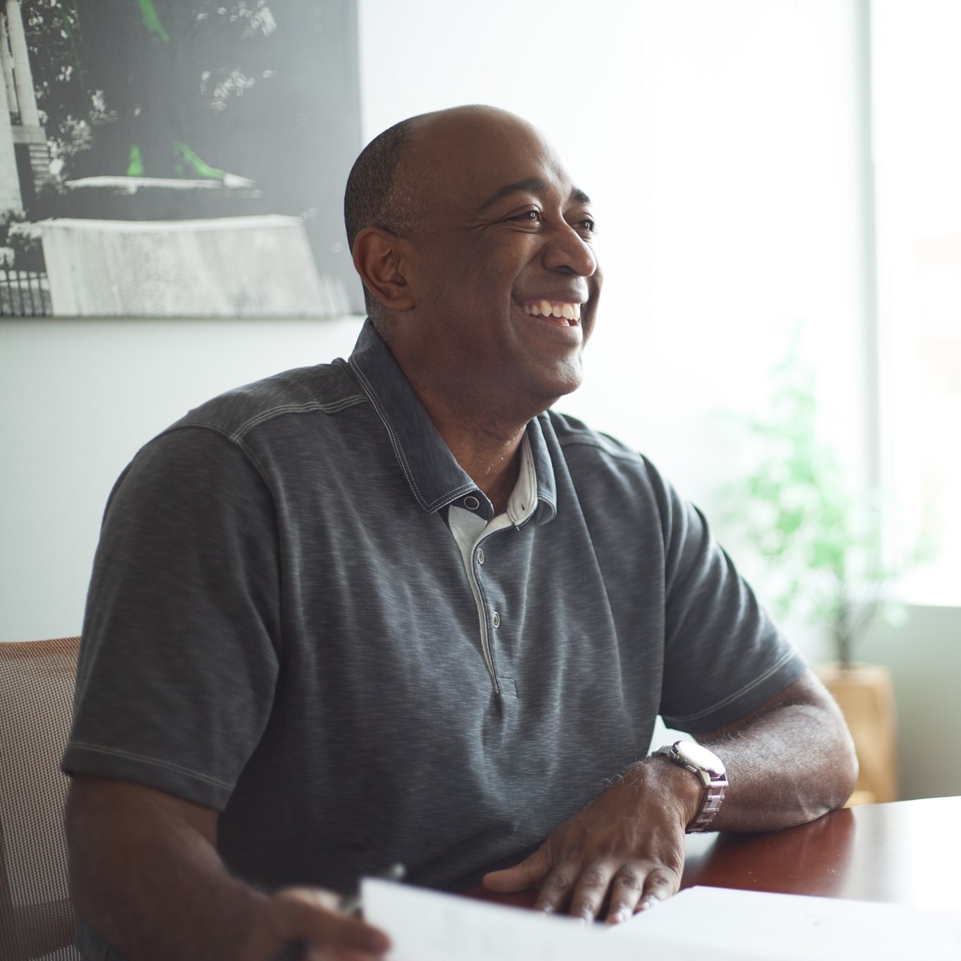 Smiling man sitting at a table with a window and greenery in the background.
