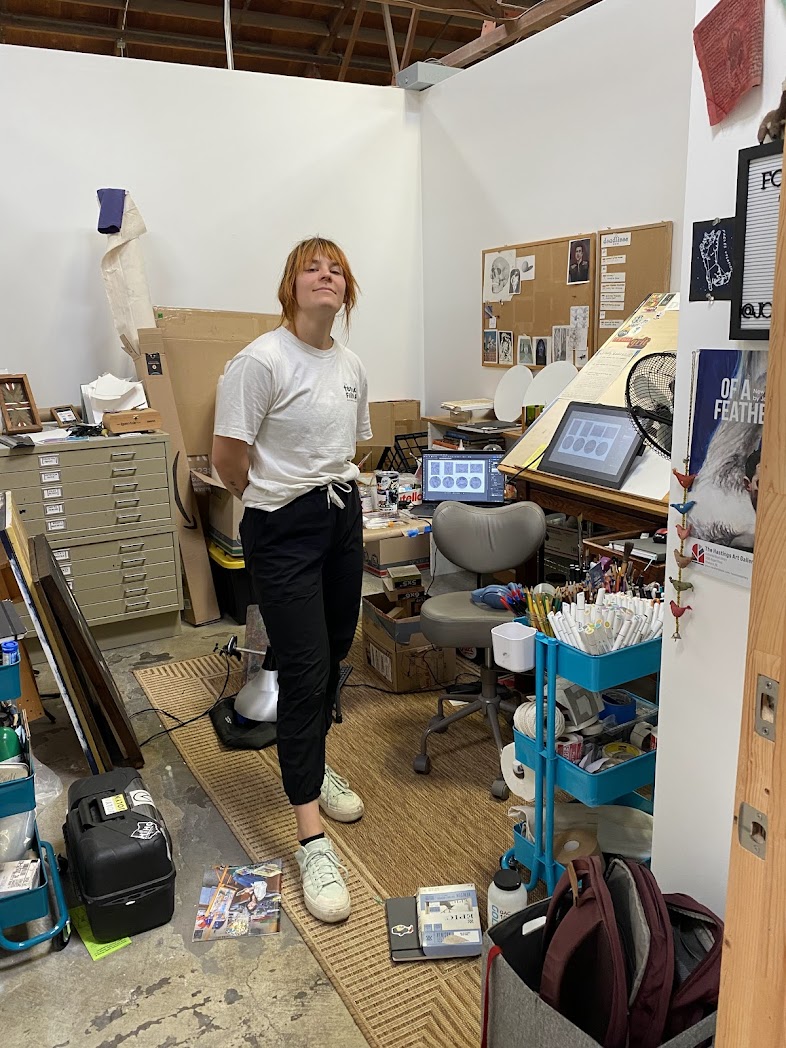 Young woman with red hair in a white t-shirt and black pants stands in a cluttered workspace with art supplies and equipment.