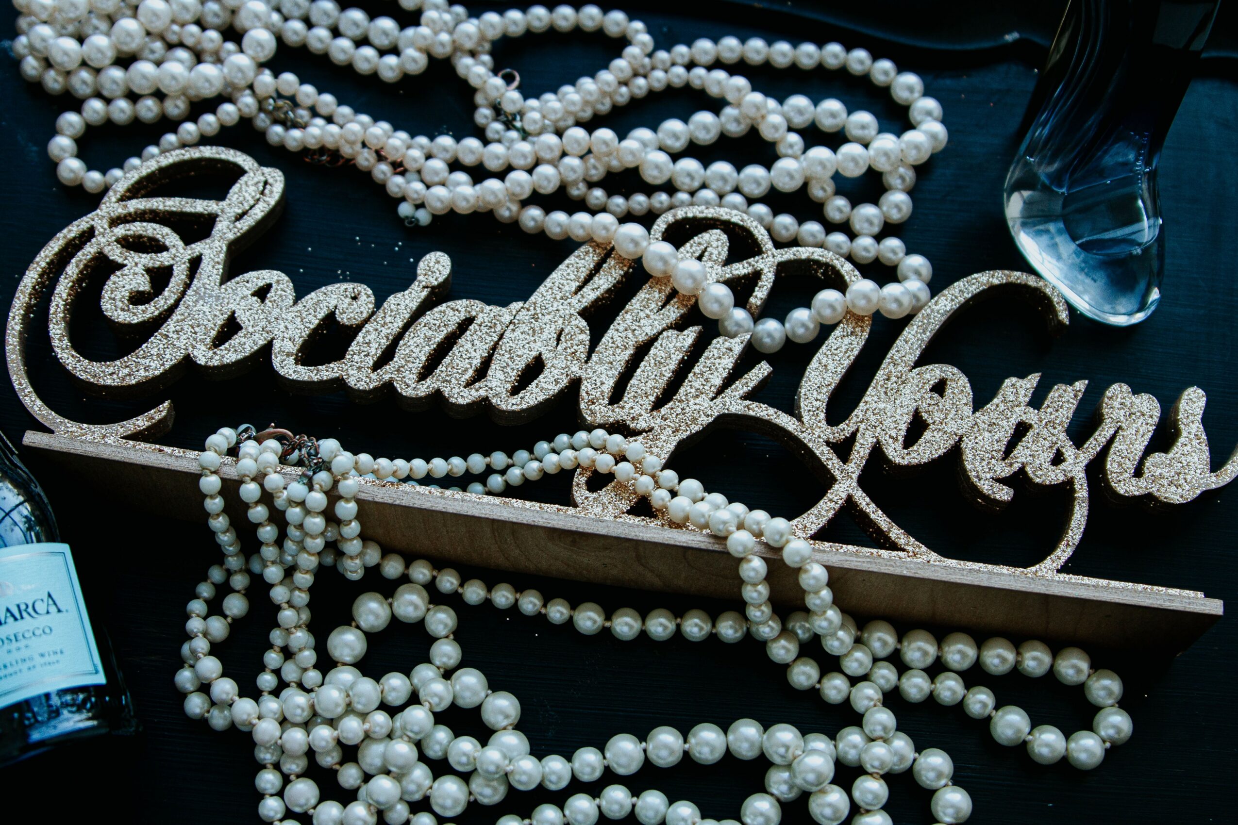 Decorative sign with cursive text, surrounded by pearl necklaces, a pen, and bottles, on a dark surface.