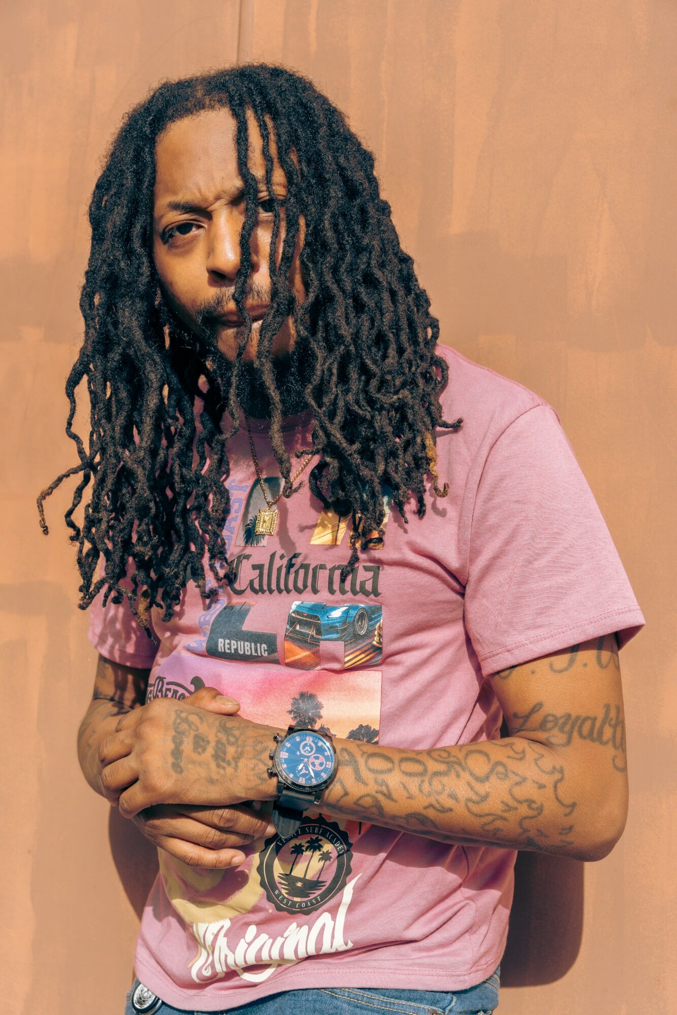 Person with long dreadlocks wearing a pink graphic T-shirt, standing against a wooden background.