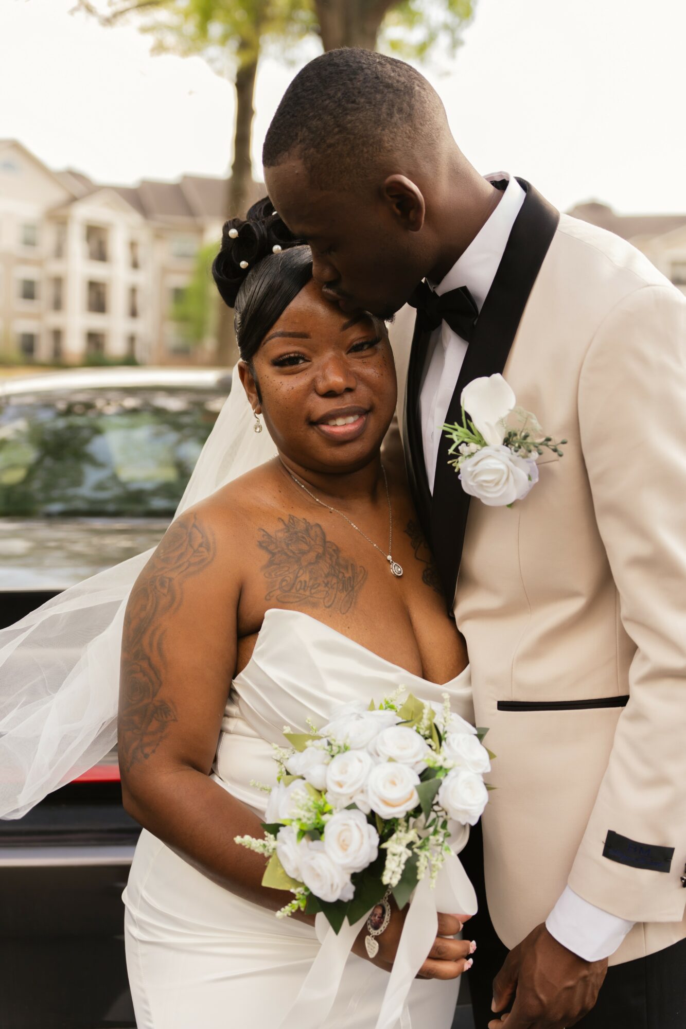 A bride and groom share a moment outdoors, the groom kissing the bride's forehead, she holds a bouquet, both in wedding attire.