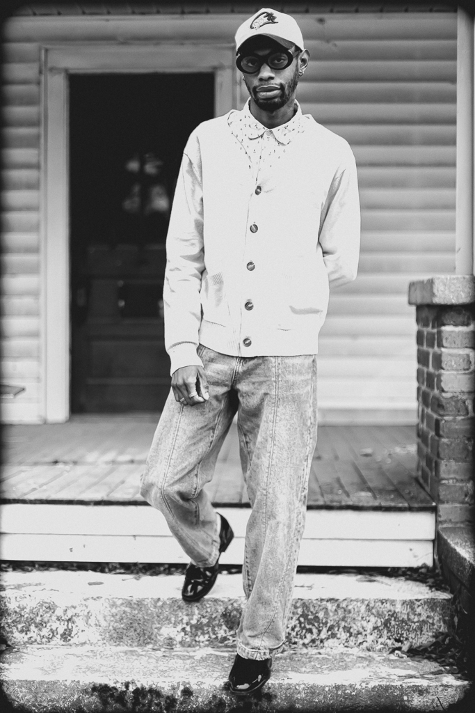 Man standing on steps in front of a building, wearing sunglasses, a cap, a light jacket, and loose pants.