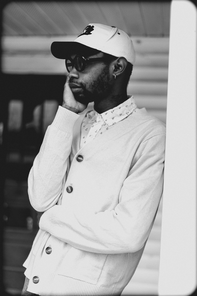 Man wearing a cap, sunglasses, and a buttoned cardigan, standing outdoors with a thoughtful expression.
