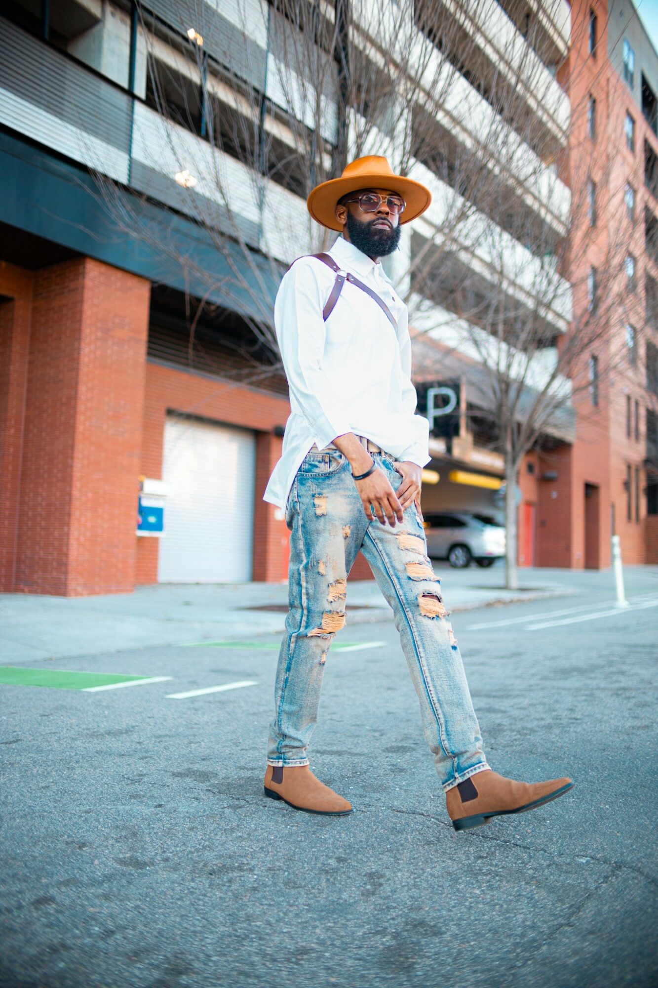Person walking on city street wearing a wide-brimmed hat, white shirt, ripped jeans, and tan shoes, with buildings in background.