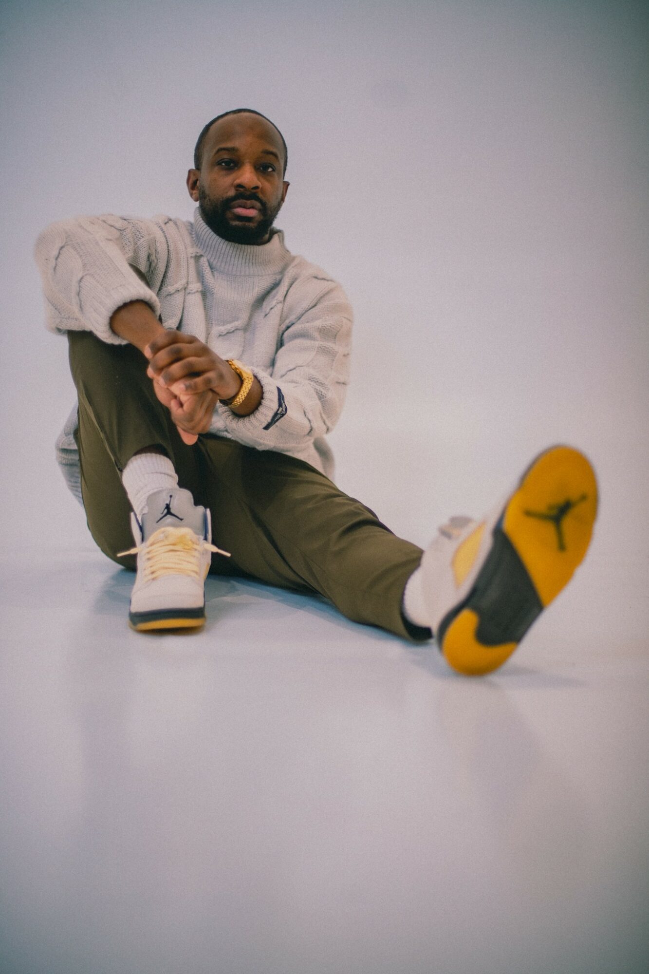 Person sitting on the floor with legs extended, wearing a light sweater and sneakers, against a plain background.