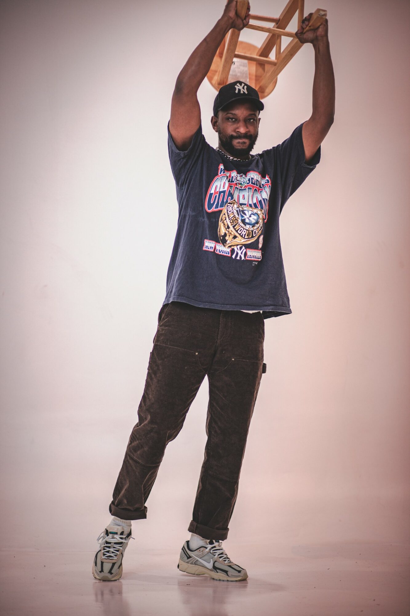 Person with a beard and cap holding a small wooden chair above head, smiling, standing against plain background.