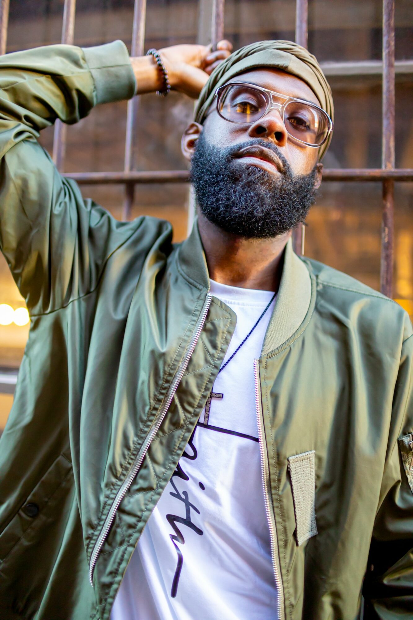 Man with glasses and a beard, wearing a green jacket and a cap, leaning against a metal fence.