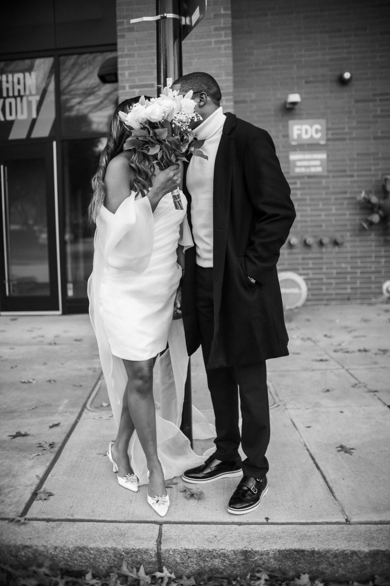 Two people stand close together on a sidewalk, one holding a large bouquet of flowers in front of their face, in front of a brick building.