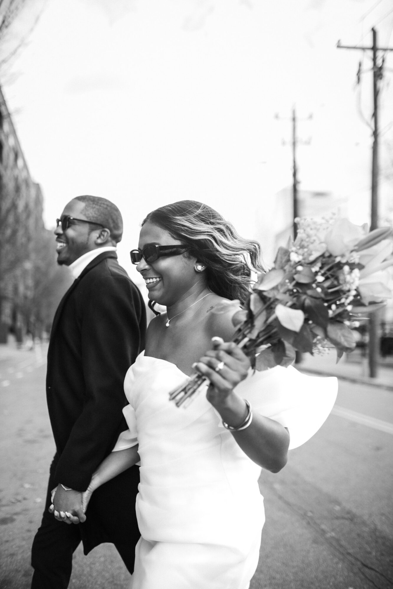 Two people walking outdoors, one holding a bouquet of flowers, both smiling and wearing sunglasses, with power lines in the background.