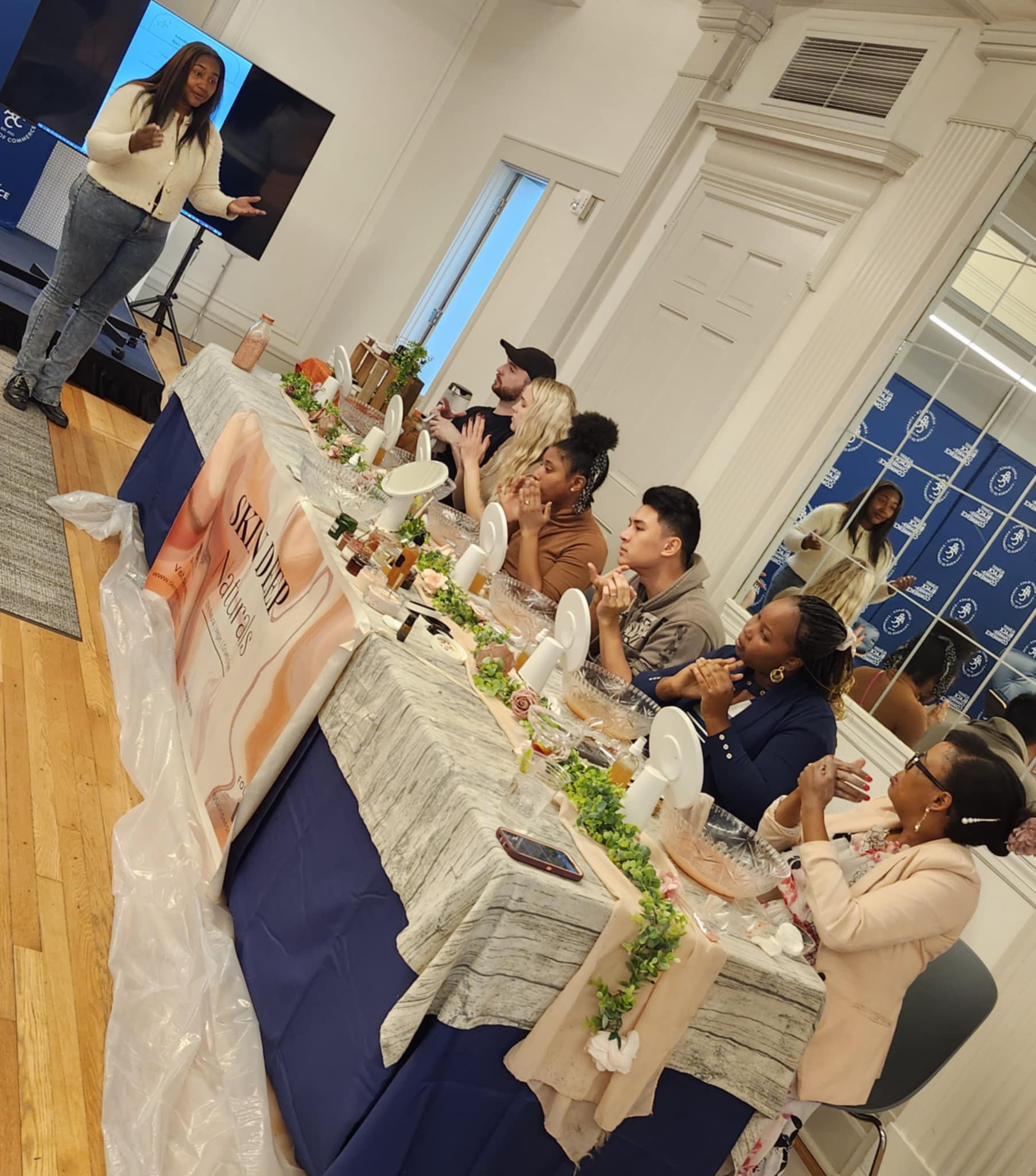 Woman presenting to seated group at a long table in a bright room with white walls and large windows.