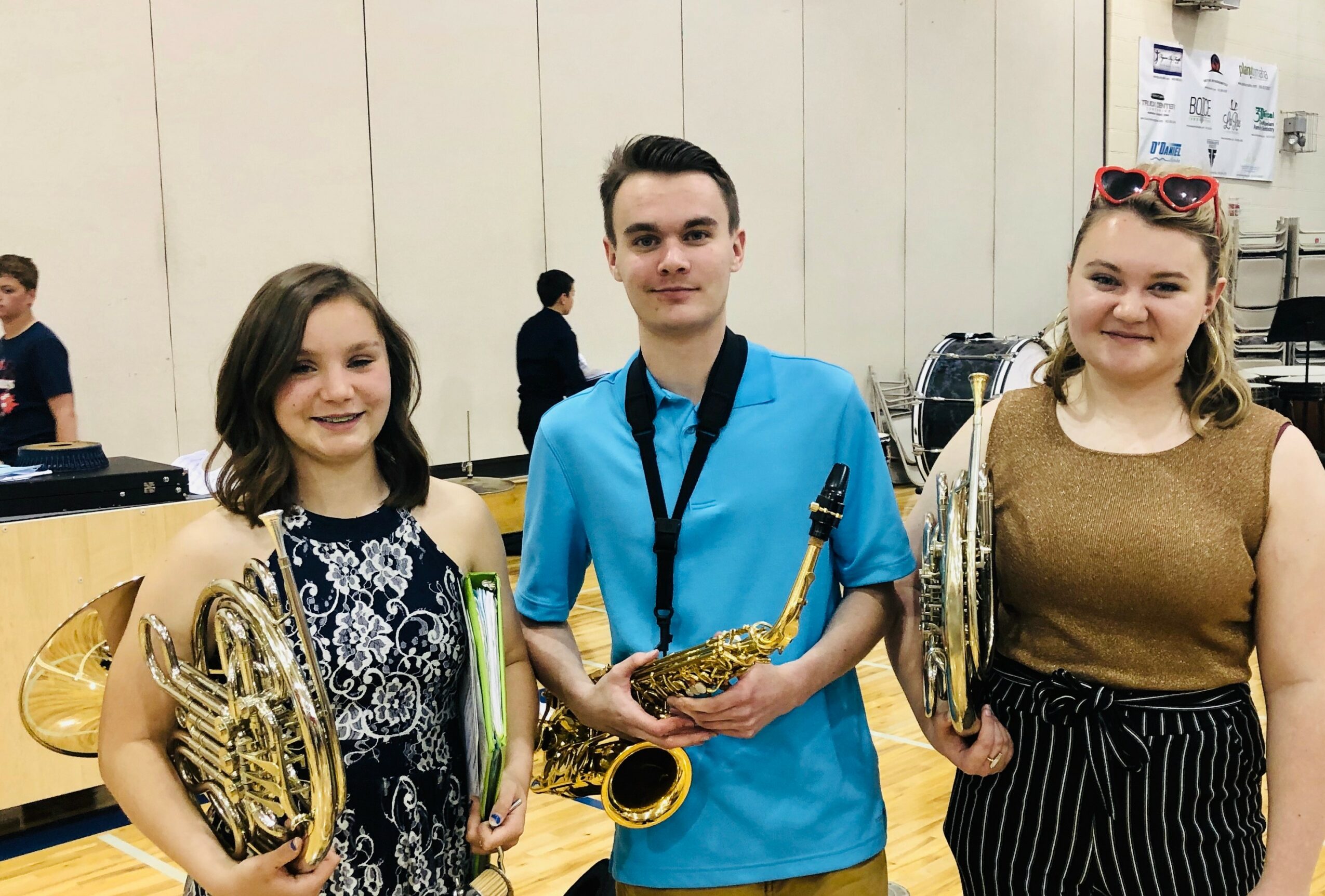 Three people holding musical instruments in a room with a beige wall and other people in the background.