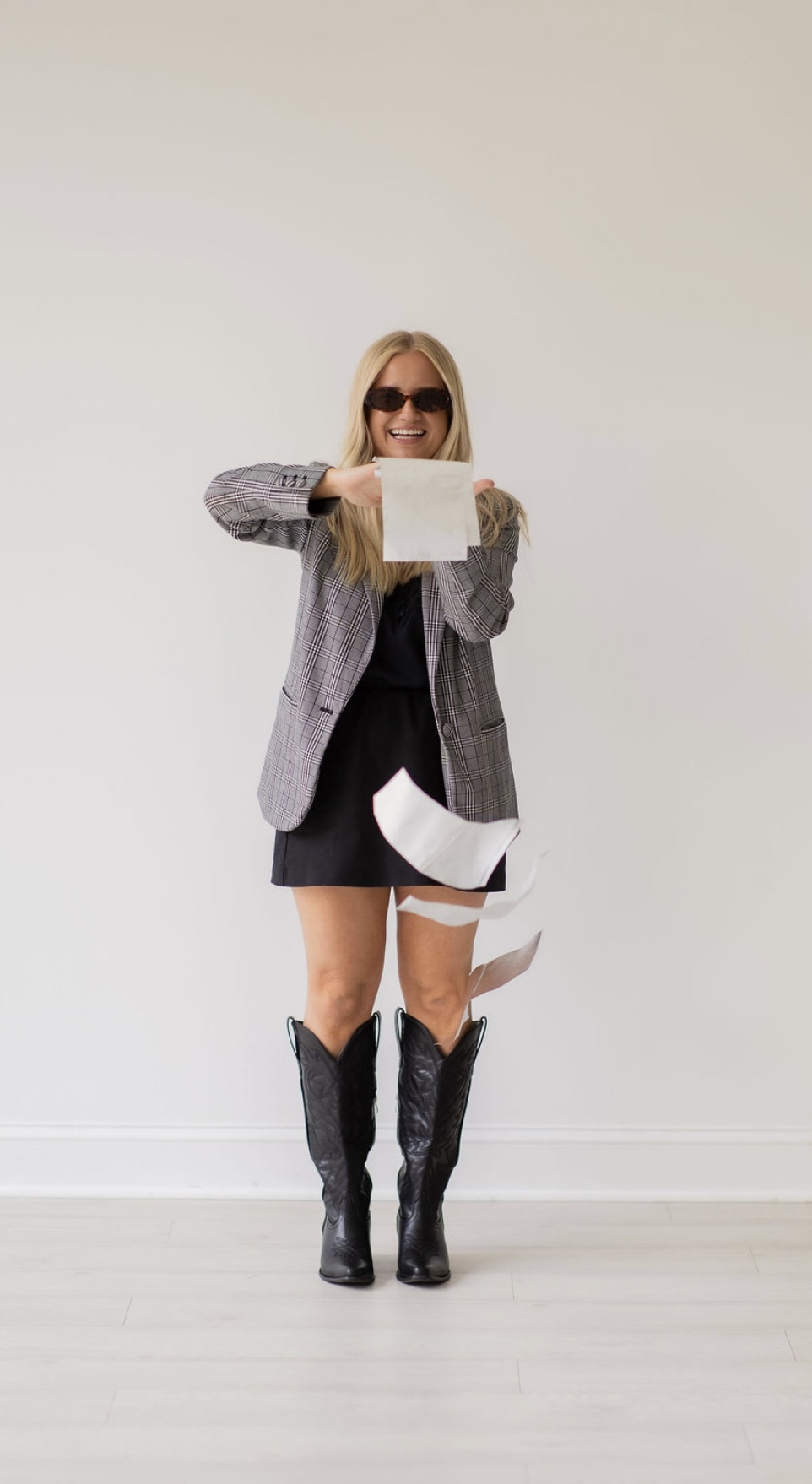 Woman with sunglasses holding a paper, standing against a plain wall, wearing a blazer, skirt, and boots.