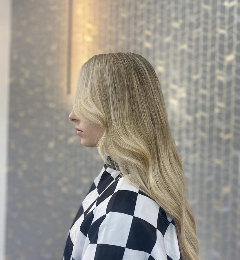 Side view of a woman with long, wavy blonde hair wearing a checkered shirt, standing against a textured wall.