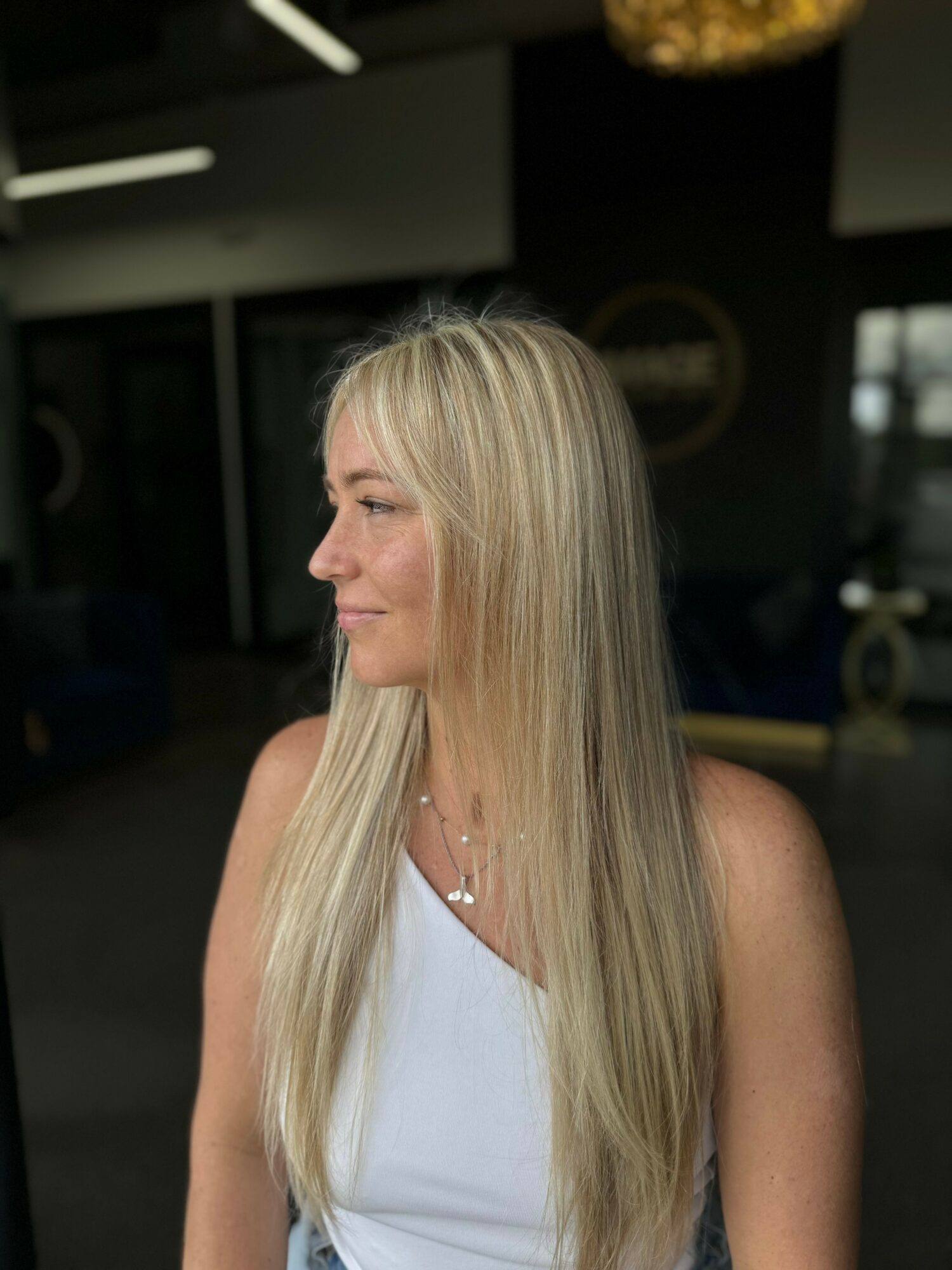 Side profile of a woman with long blonde hair wearing a white top, indoors with dark background.