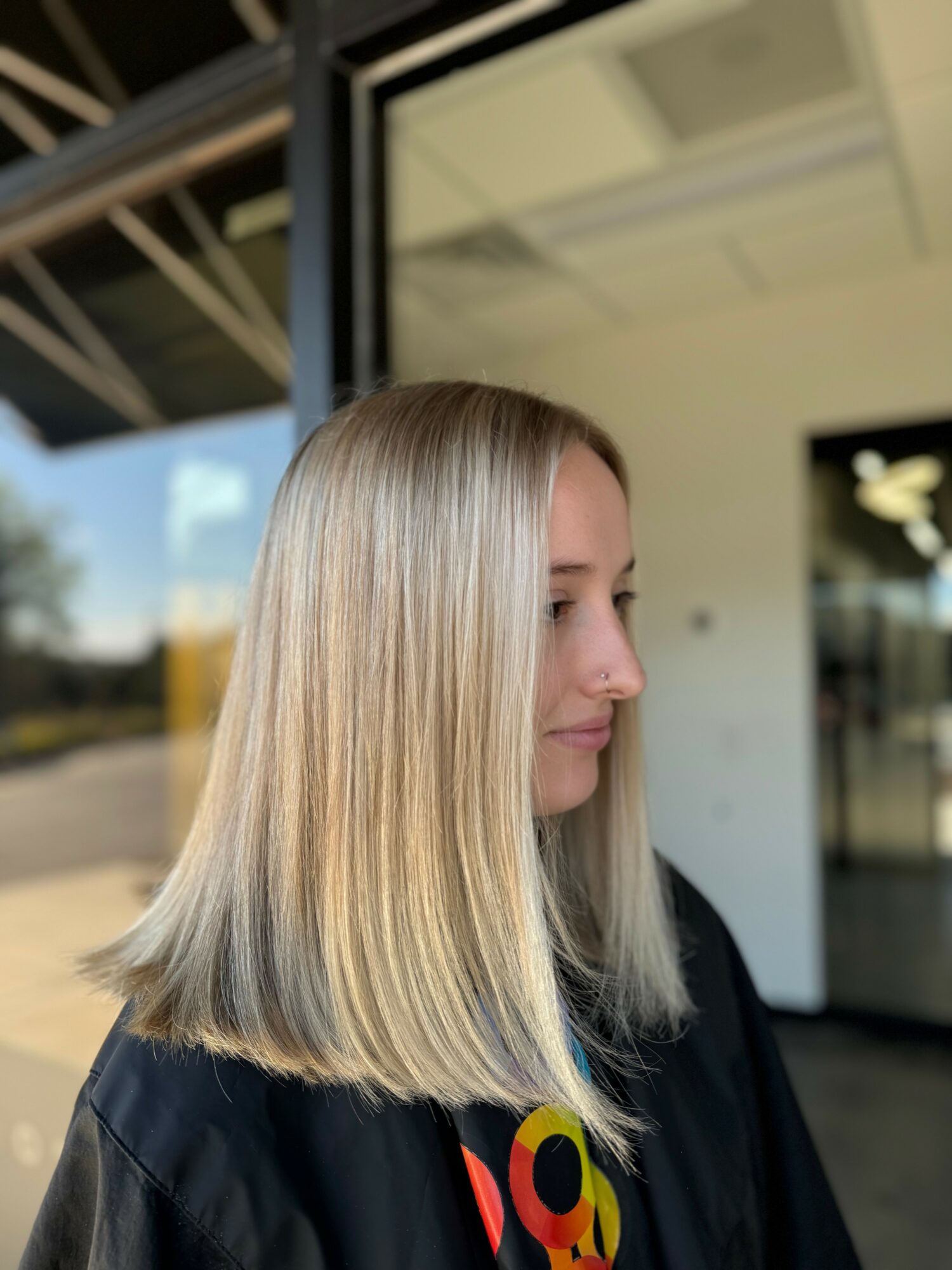 Side profile of a woman with straight, blonde hair standing indoors near a glass door.