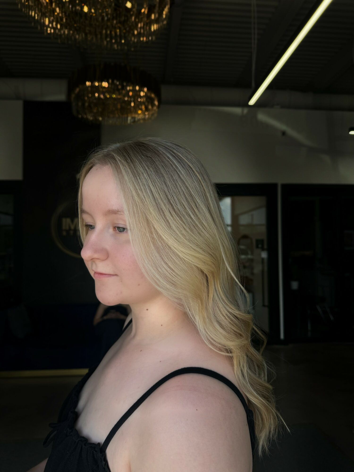 Side profile of a woman with blonde hair and black straps, indoors with ceiling lights and chandeliers visible.