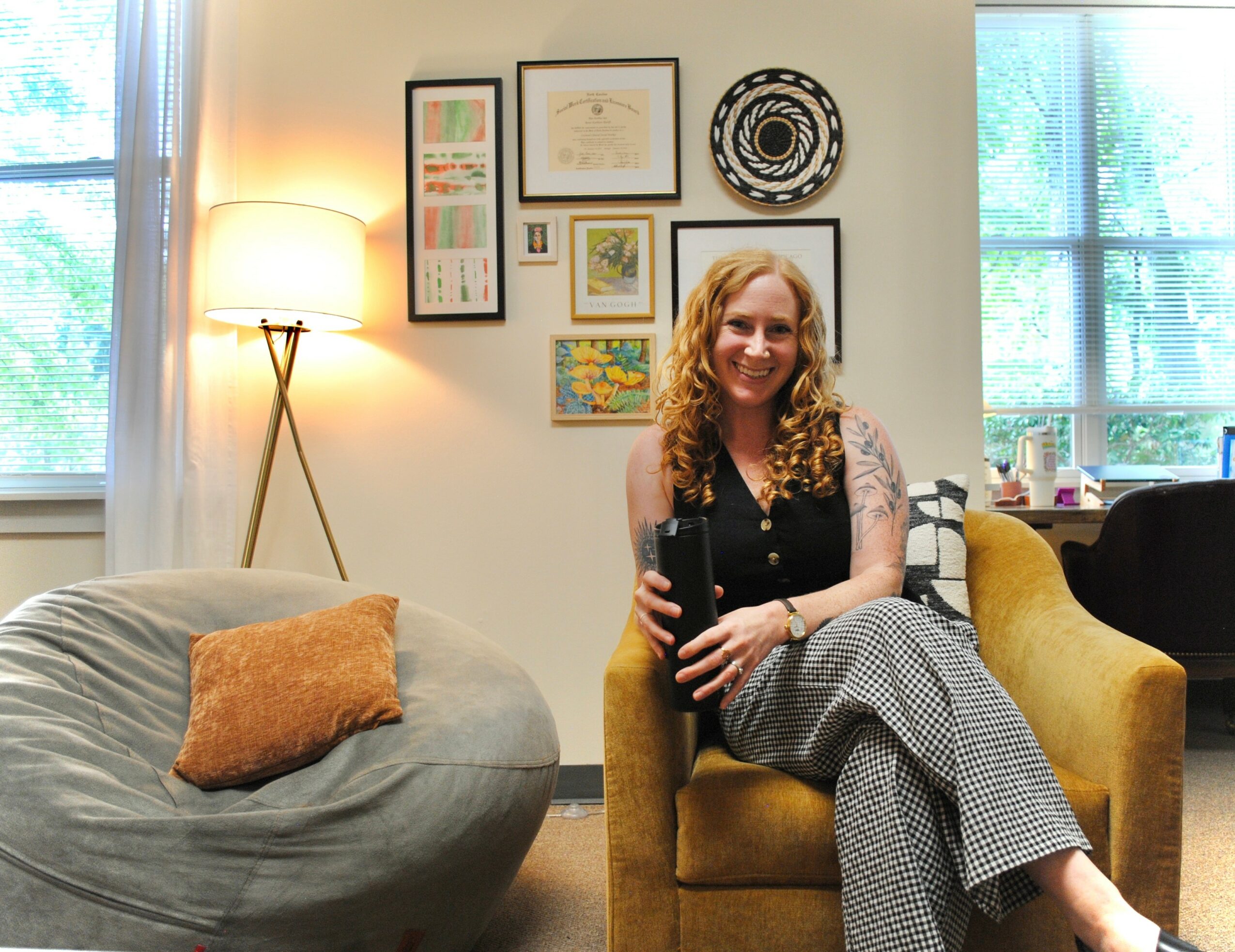 Woman with curly hair sitting on a yellow armchair in a living room, smiling, with framed art on the wall behind her.