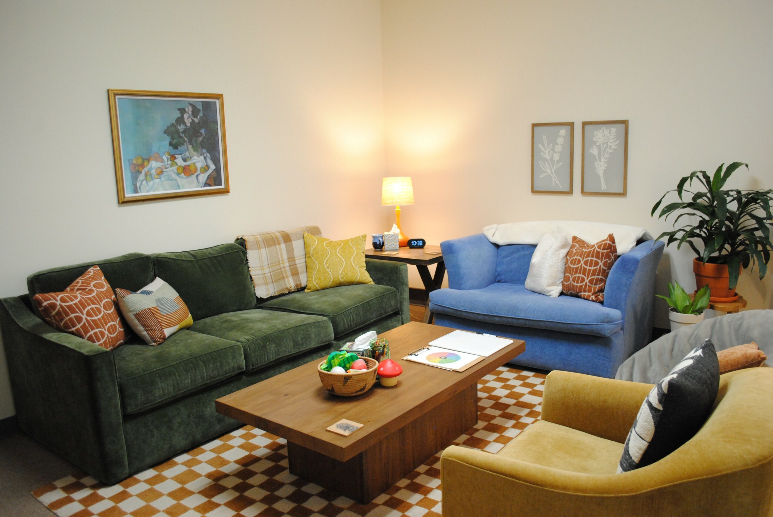 Living room with green and blue sofas, a wooden coffee table, framed artwork, and a potted plant, illuminated by a lamp.