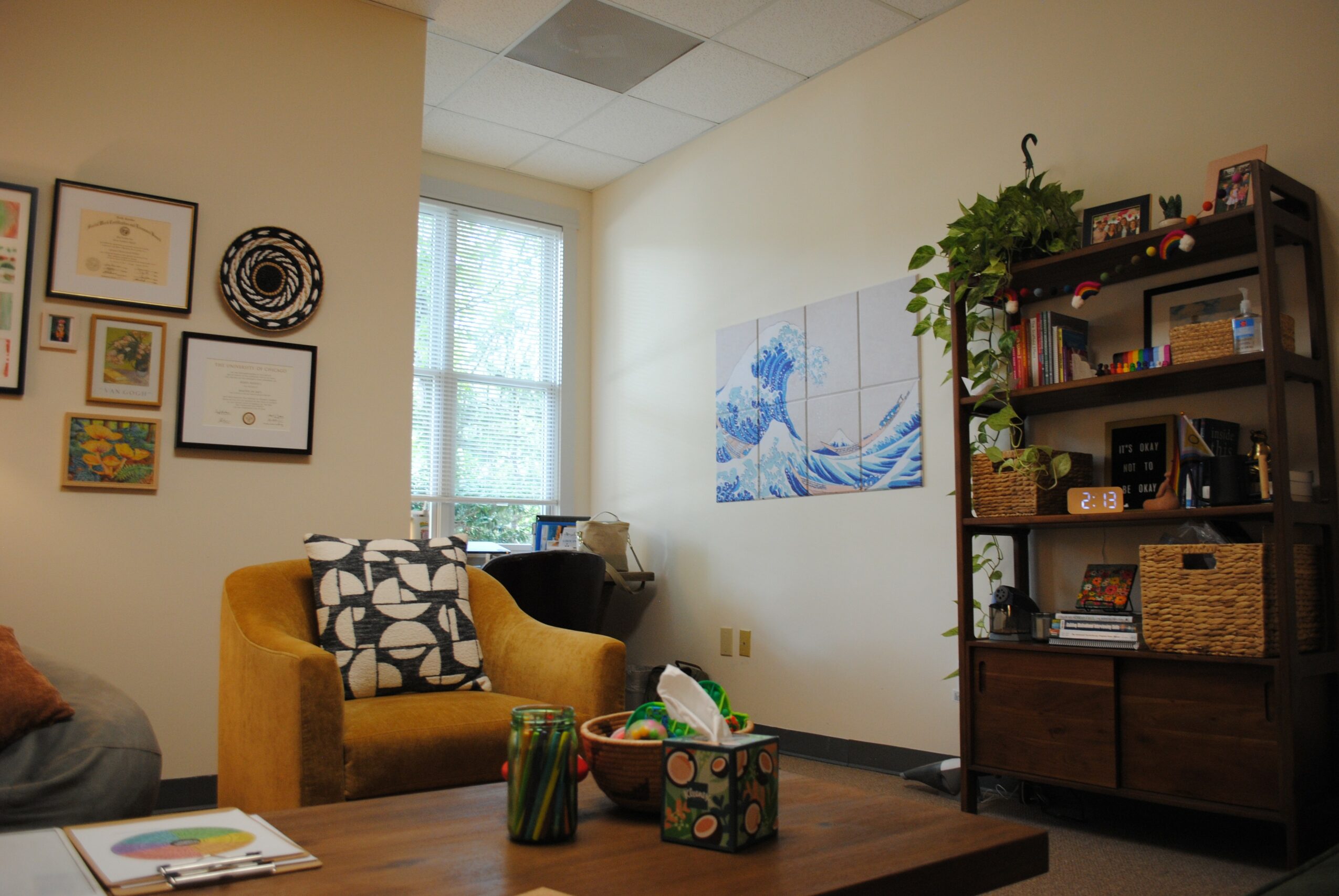 Living room with yellow armchair, bookshelf, wall art, and window, decorated with plants and framed pictures.