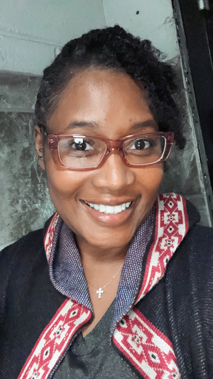 Woman with short curly hair, glasses, smiling, wearing a patterned scarf and a cross necklace, indoors.
