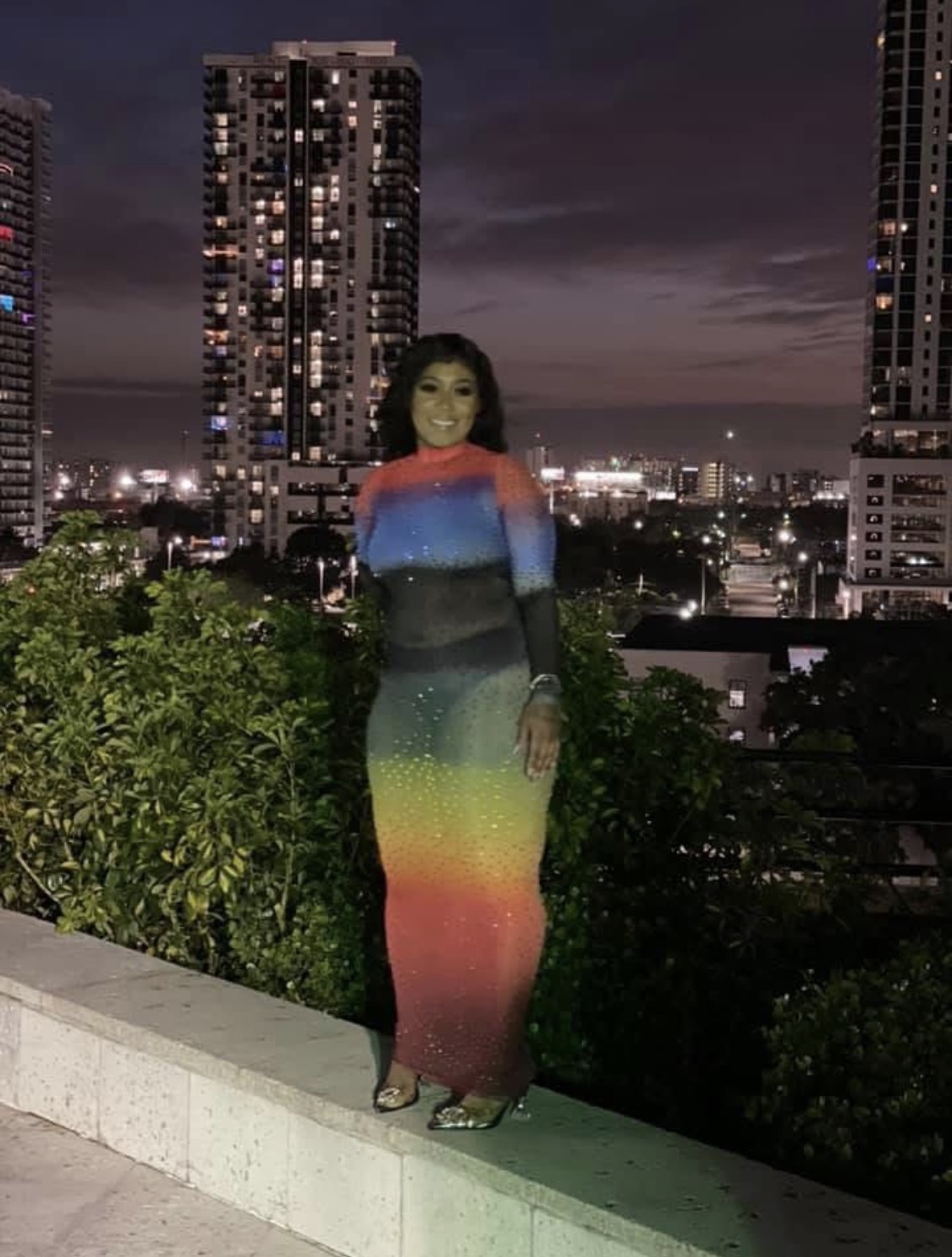 Woman in a colorful, rainbow-patterned dress standing outdoors at night with city buildings in background.