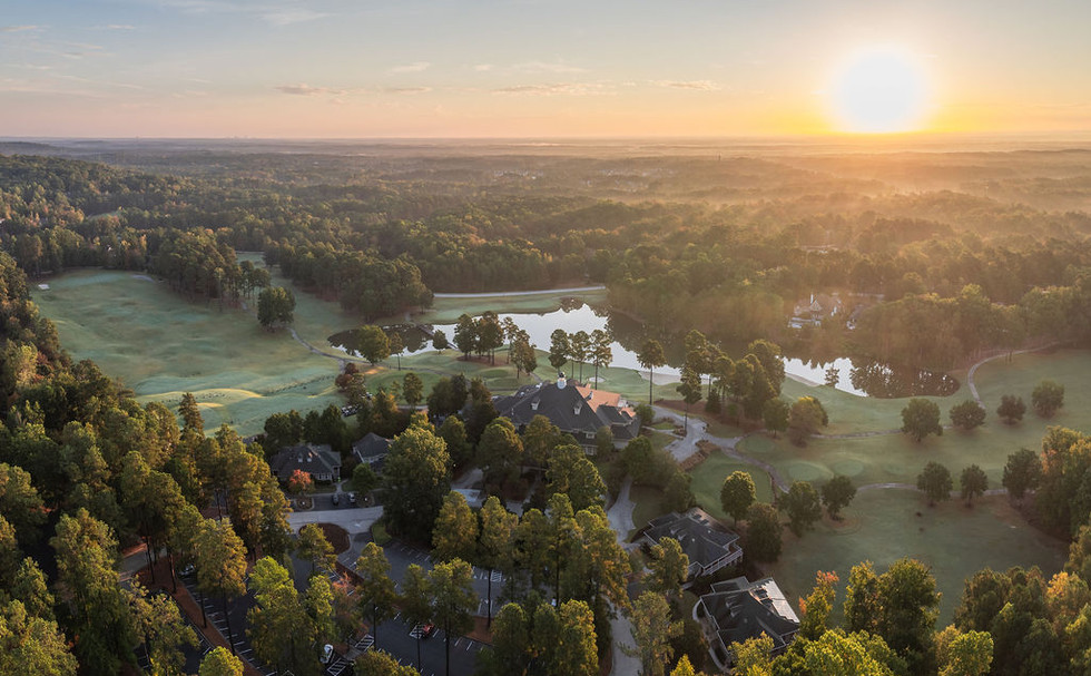 Sunset over a landscape with trees, water, and houses, with a clear sky and horizon in the distance.