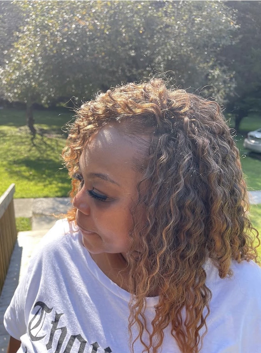 Woman with curly hair outdoors, sunlight, trees, and a car in the background, wearing a white shirt.