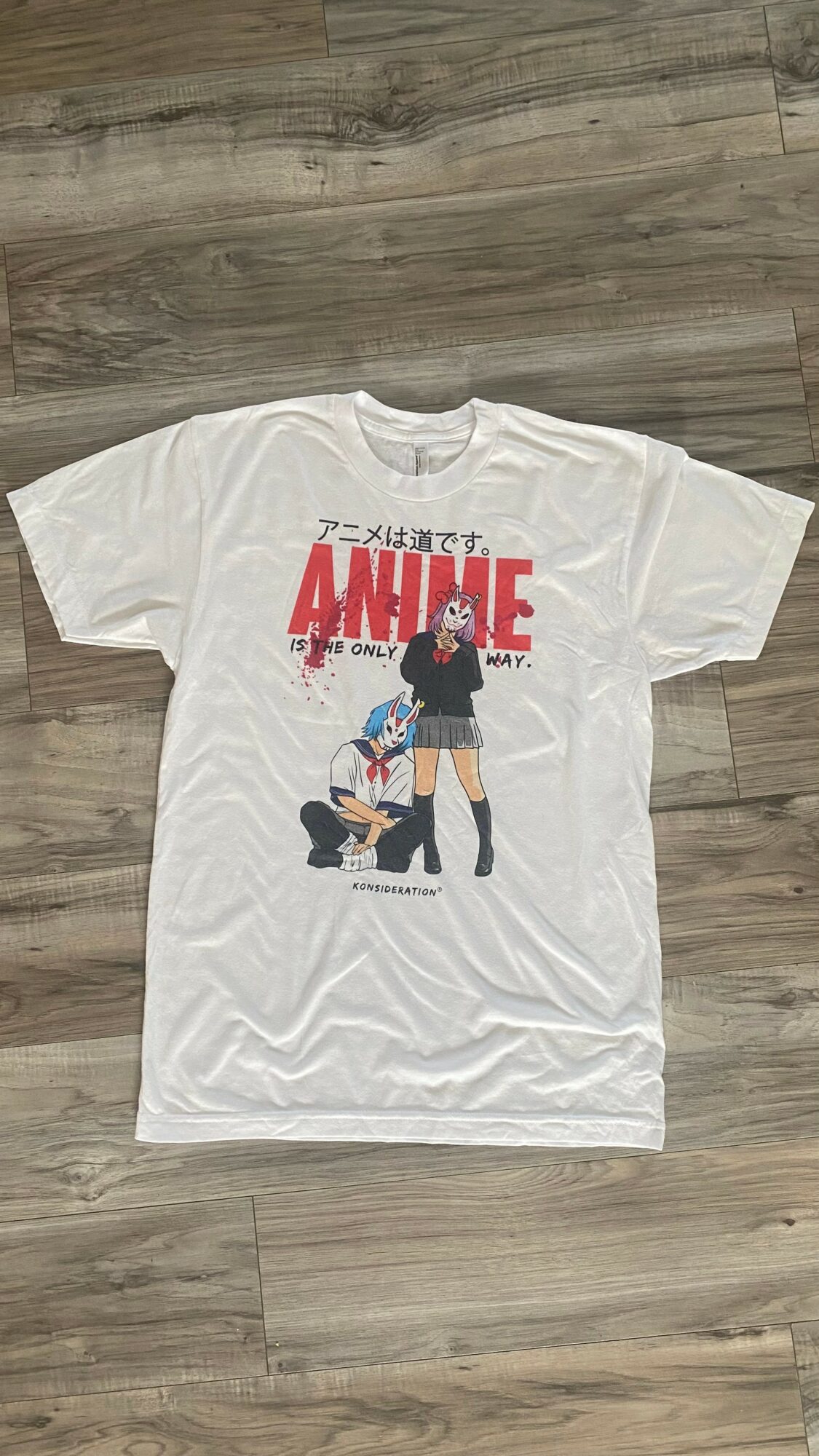 White T-shirt with anime-style illustration and red text, laid flat on a wooden floor.