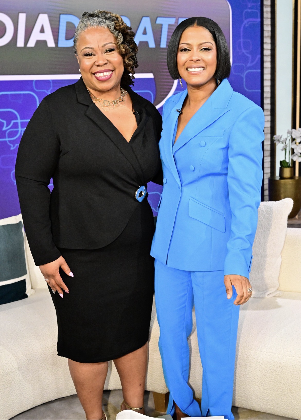 Two women stand together smiling, one in a black suit and the other in a blue suit, in a TV studio setting.