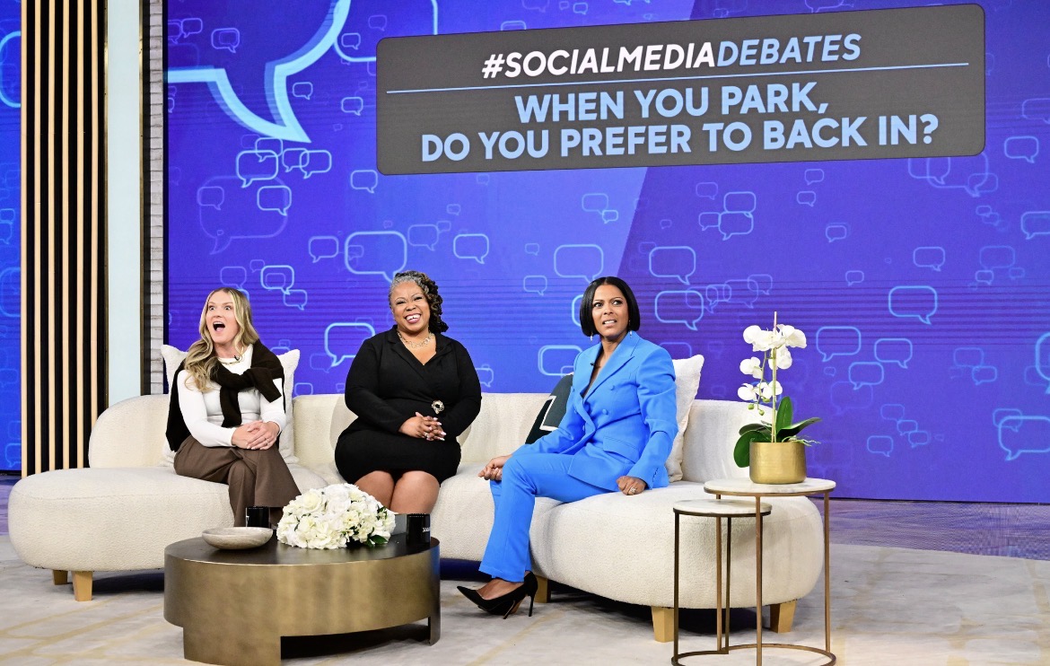 Three women sit on a stage with a blue background and social media debate question displayed, decorated with flowers and a small table.