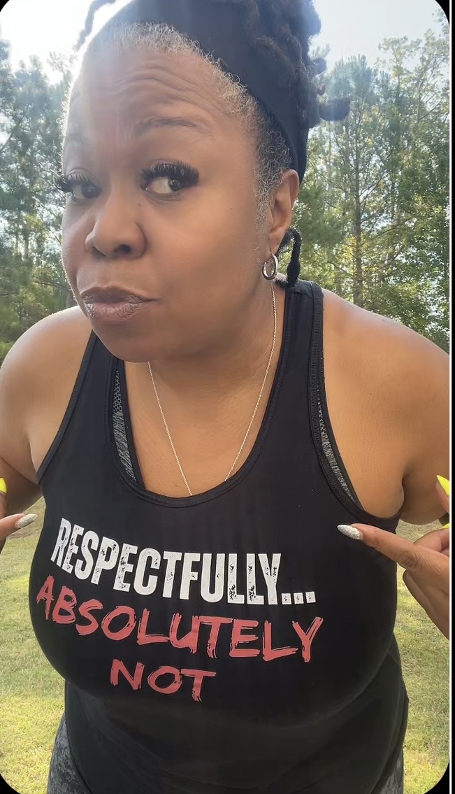 Woman outdoors wearing a black tank top with text, trees in background, looking at camera.