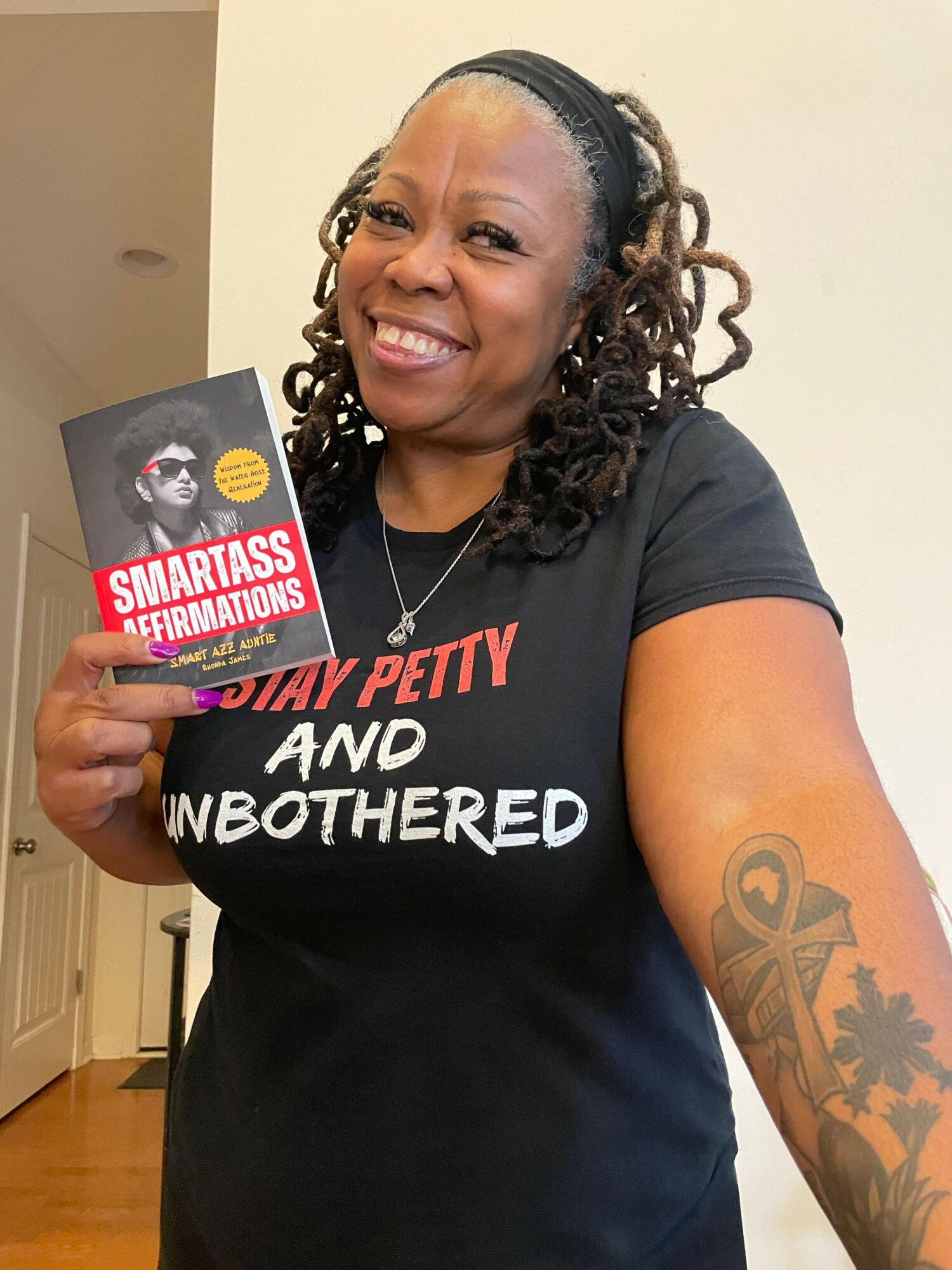 Smiling woman with dreadlocks holding a book, wearing a black T-shirt, standing indoors.