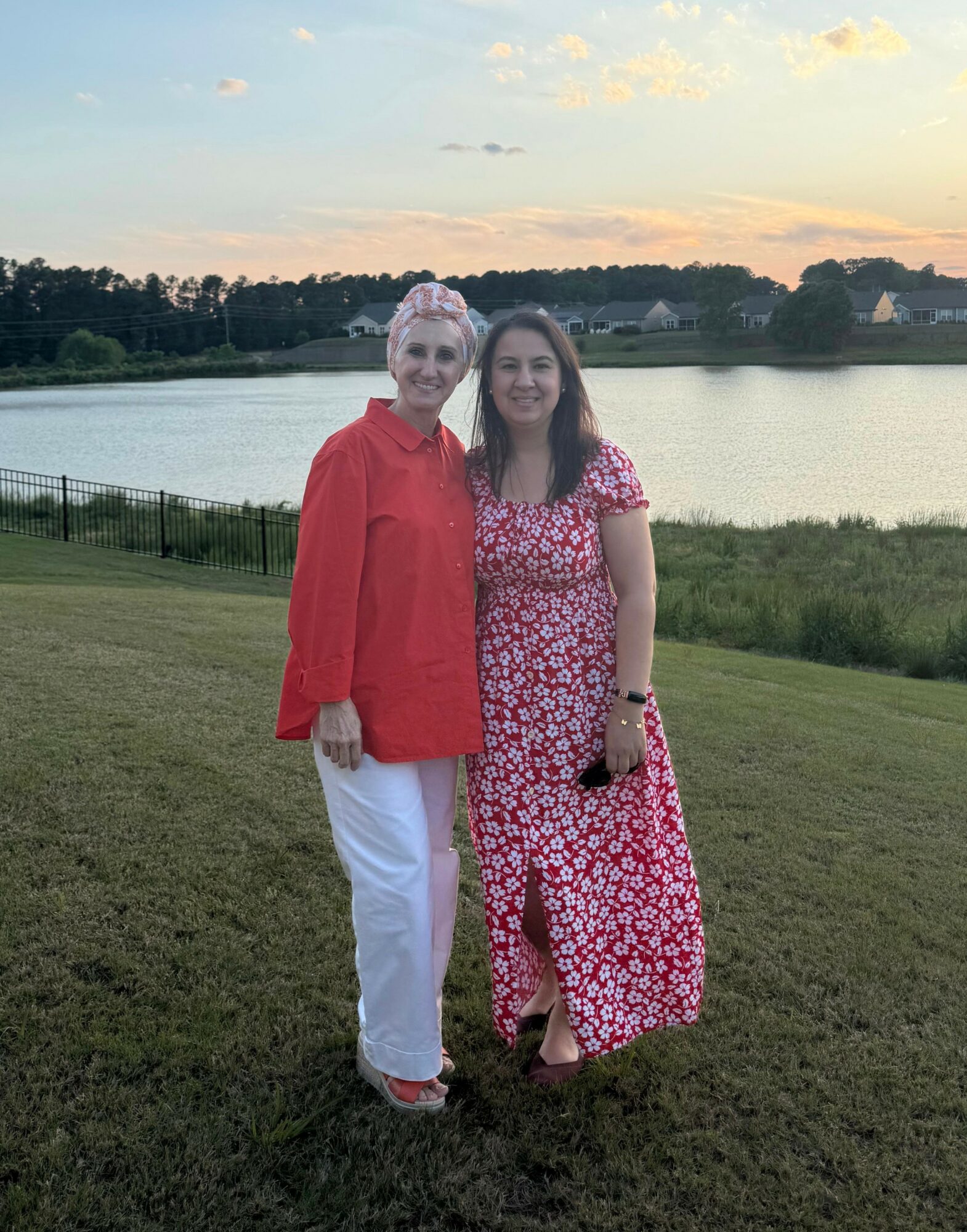 Two women standing on grass near a lake with trees and sky in the background, smiling at the camera.