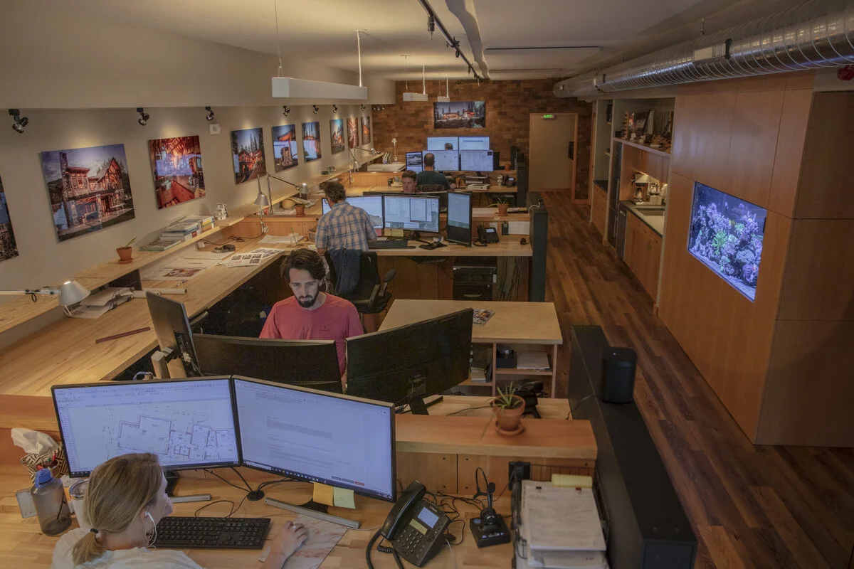 Office with multiple people working at desks with computers, monitors, and a wall-mounted TV, warm lighting, and wooden floors.
