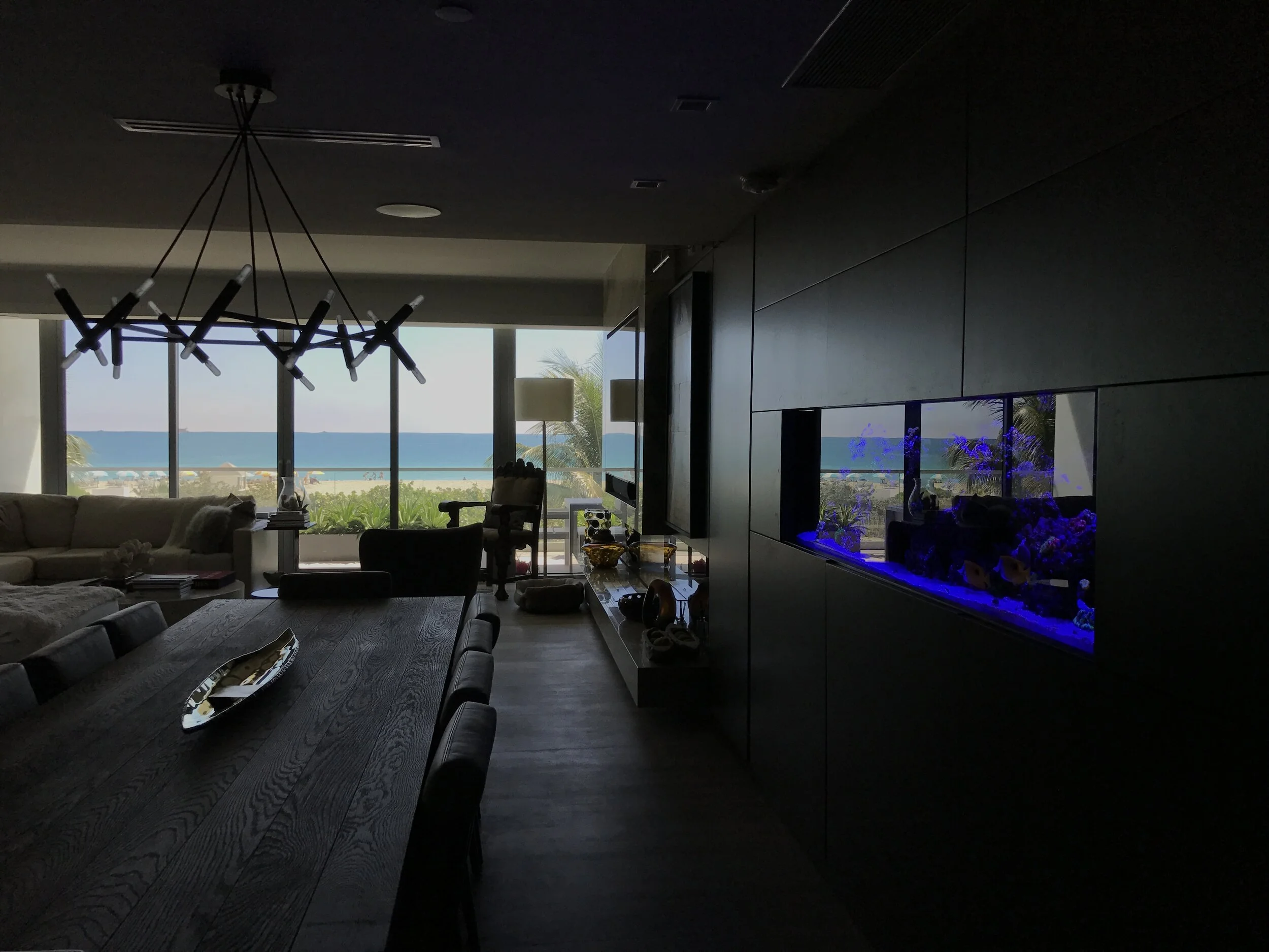 Living room with large window showing ocean view, chandelier, sofa, and a fish tank on the wall.