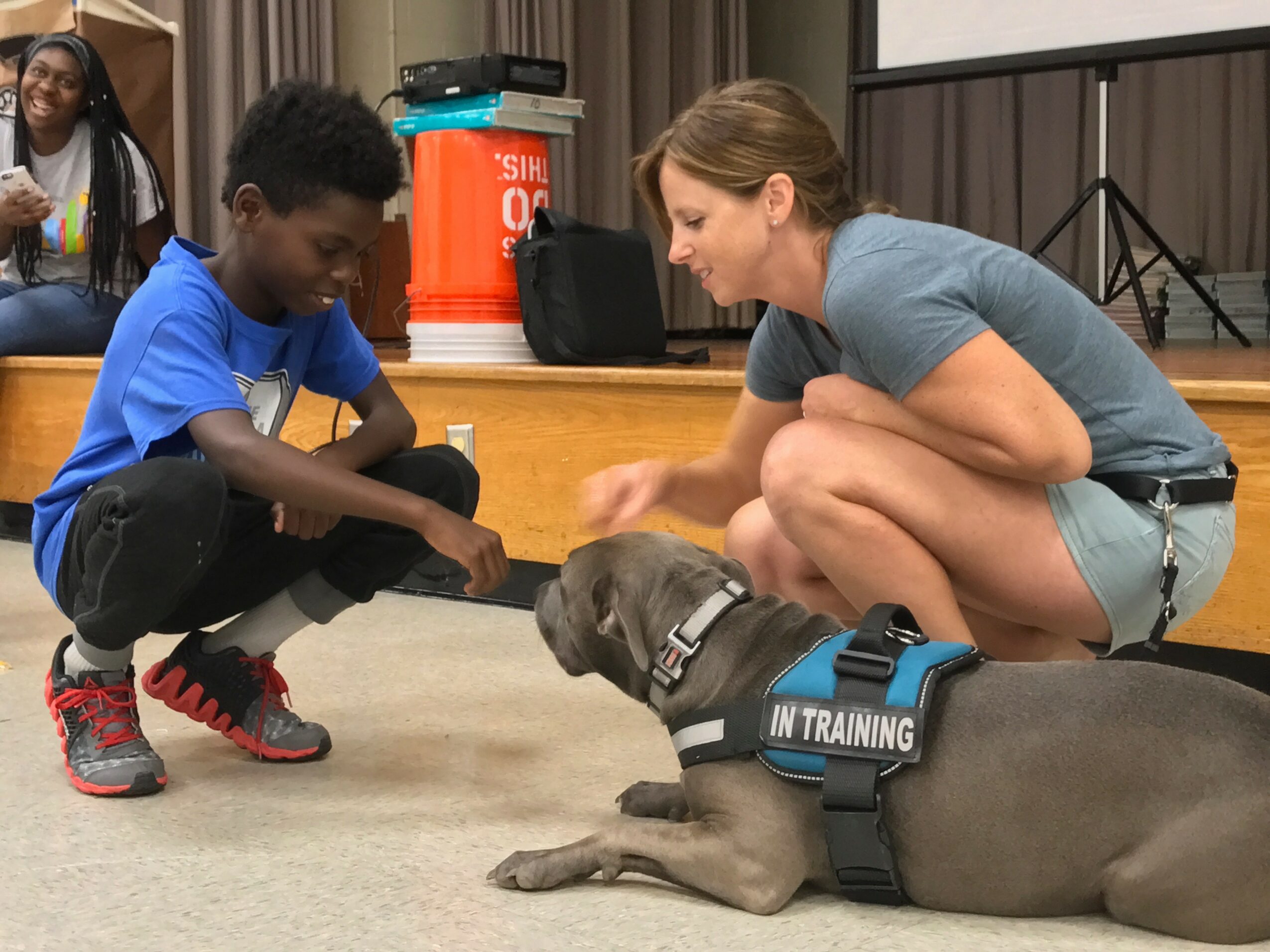Woman and child interact with a dog wearing a vest labeled 'In Training' indoors, with a stage and equipment in background.