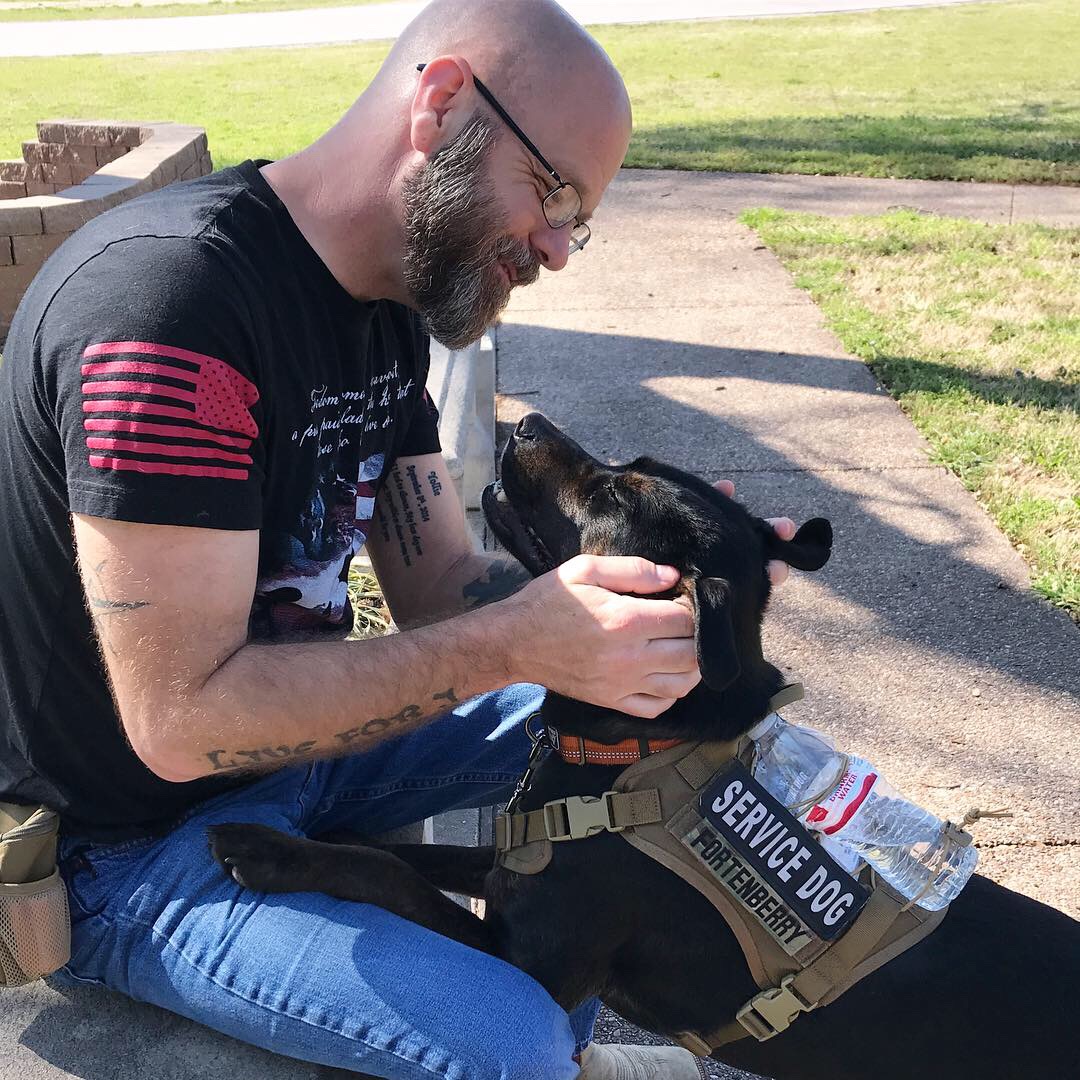 Man with glasses and beard smiling at service dog outdoors on a sunny day.