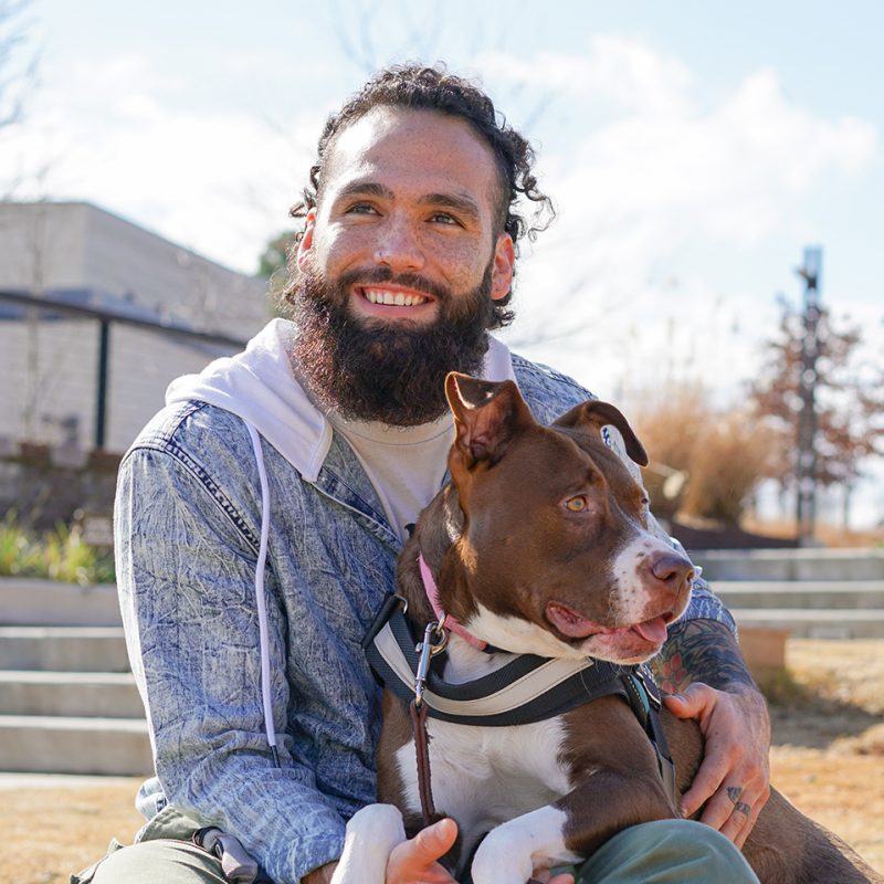 Man with a beard smiling outdoors, holding a brown and white dog wearing a harness, sunny day with trees and stairs in background.